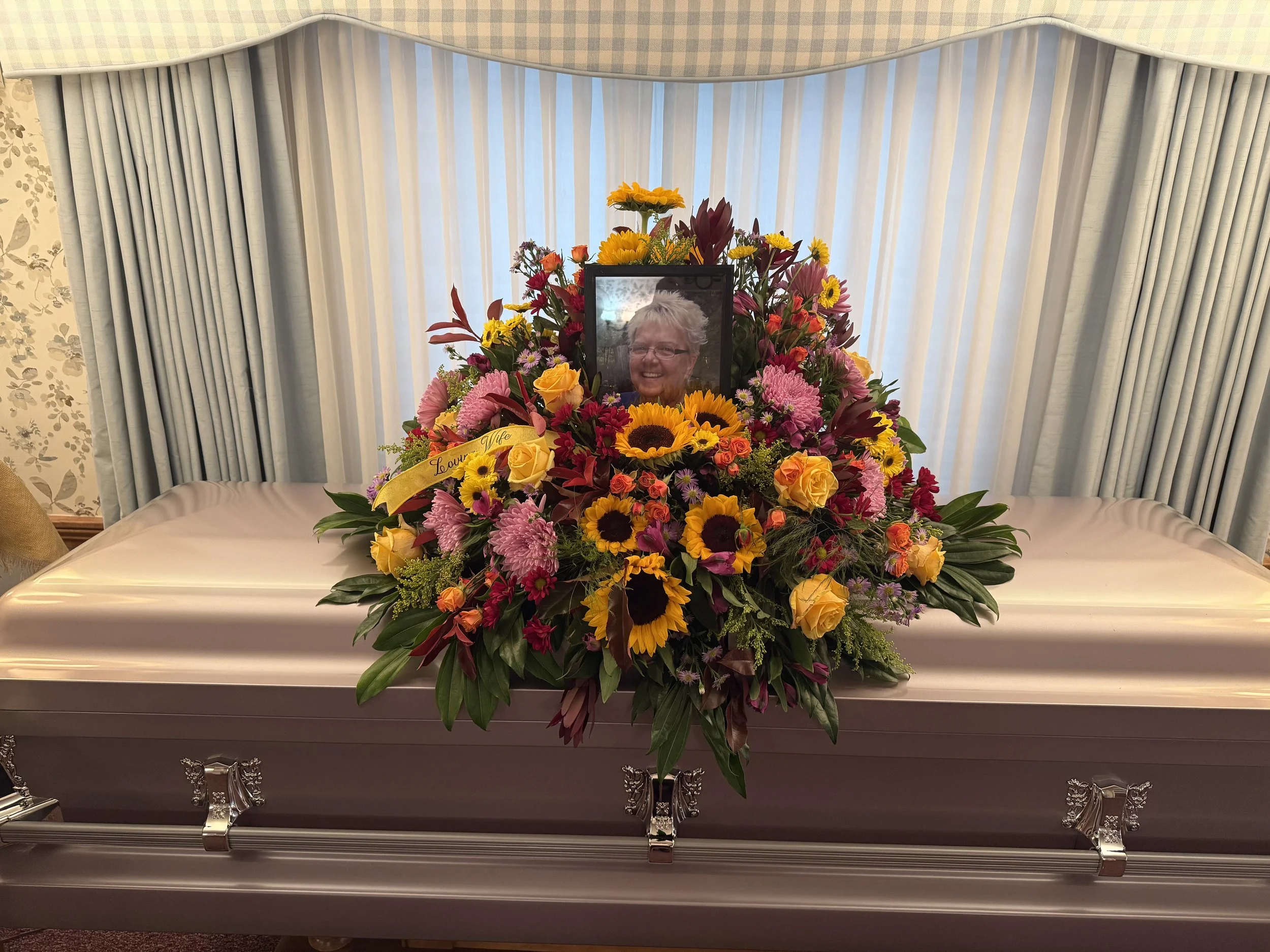 Casket with a floral arrangement and a framed photo of a smiling elderly woman on top, in a funeral home setting with curtains and floral wallpaper in the background.