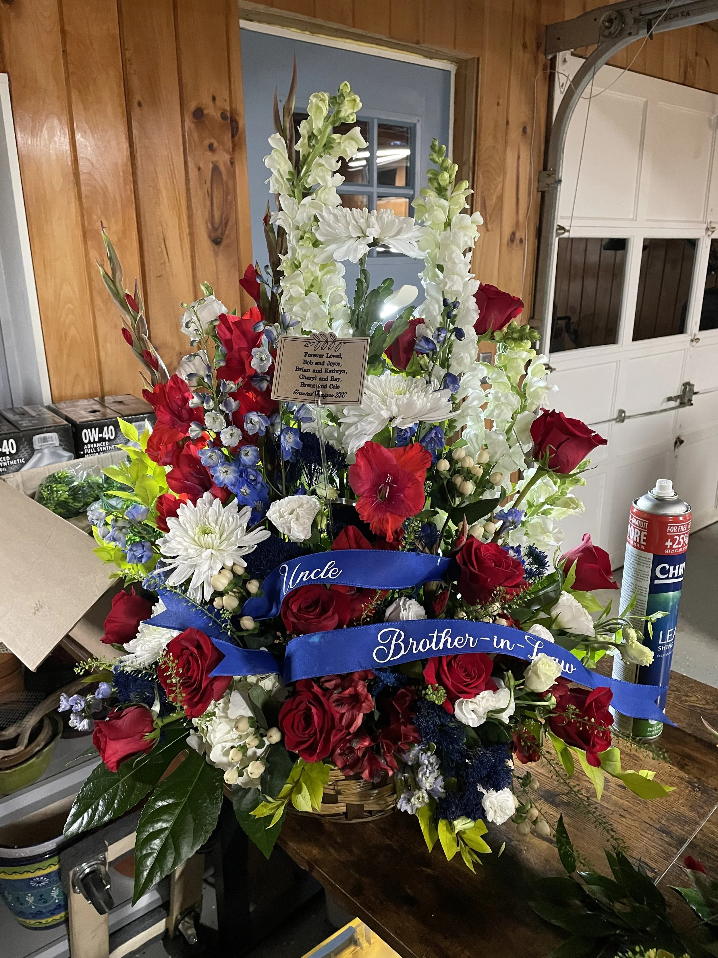 A flower arrangement with red, white, and blue flowers, including roses, in a basket. A blue ribbon with the words "Uncle" and "Brother-in-Saw" is attached. A small card with a message and a can of spray cleaner are also visible.