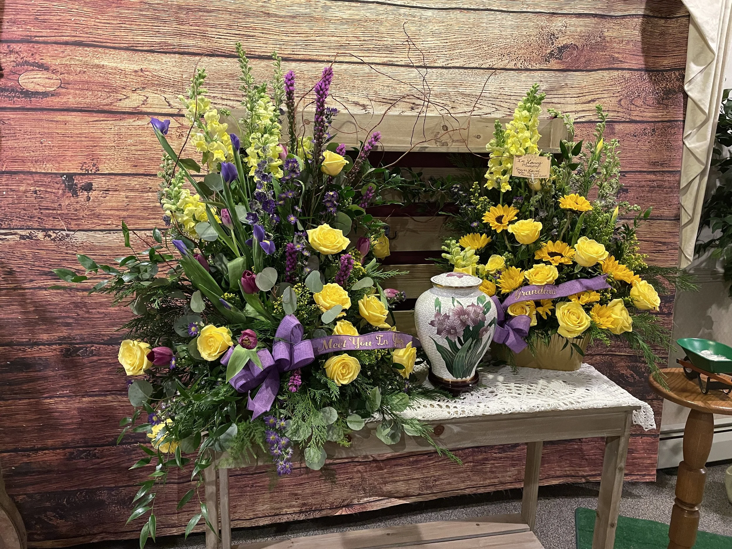 Two large flower arrangements with yellow, purple, and green flowers, placed on a small wooden table with a lace cloth, and a decorative vase in the center, against a wooden plank background.