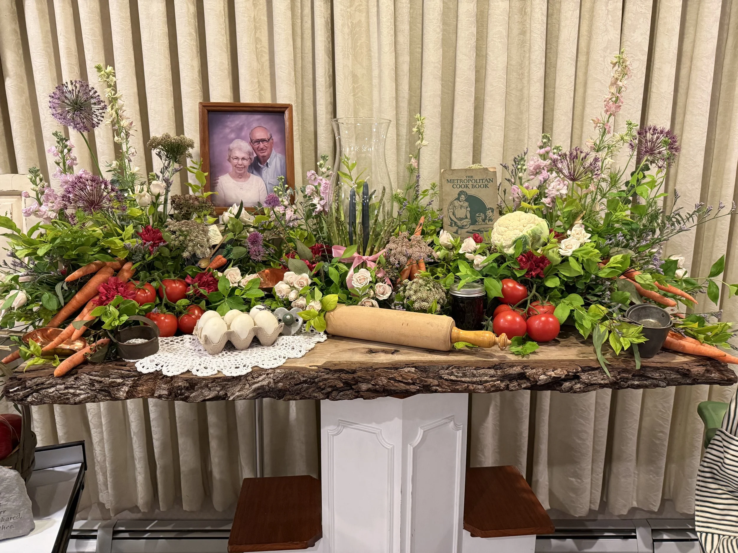 Flower arrangement with tomatoes, carrots, eggs, and various flowers on a rustic wooden table, with framed photograph and cook book in the background.