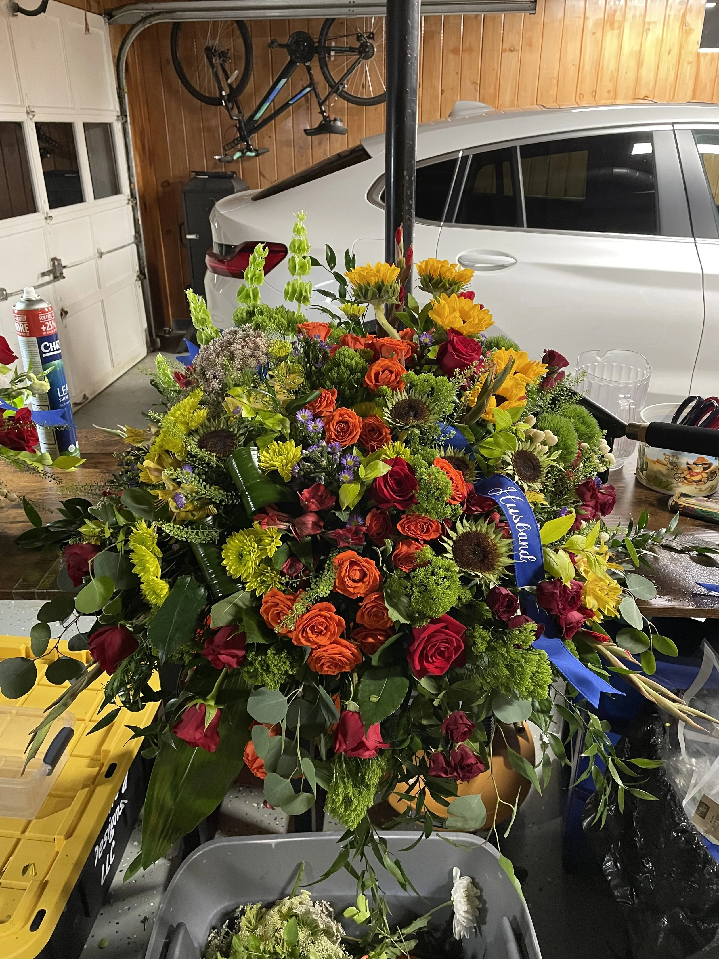 Colorful floral arrangement with roses, sunflowers, and greenery in a workshop or garage with car, bicycle, and tools in background.