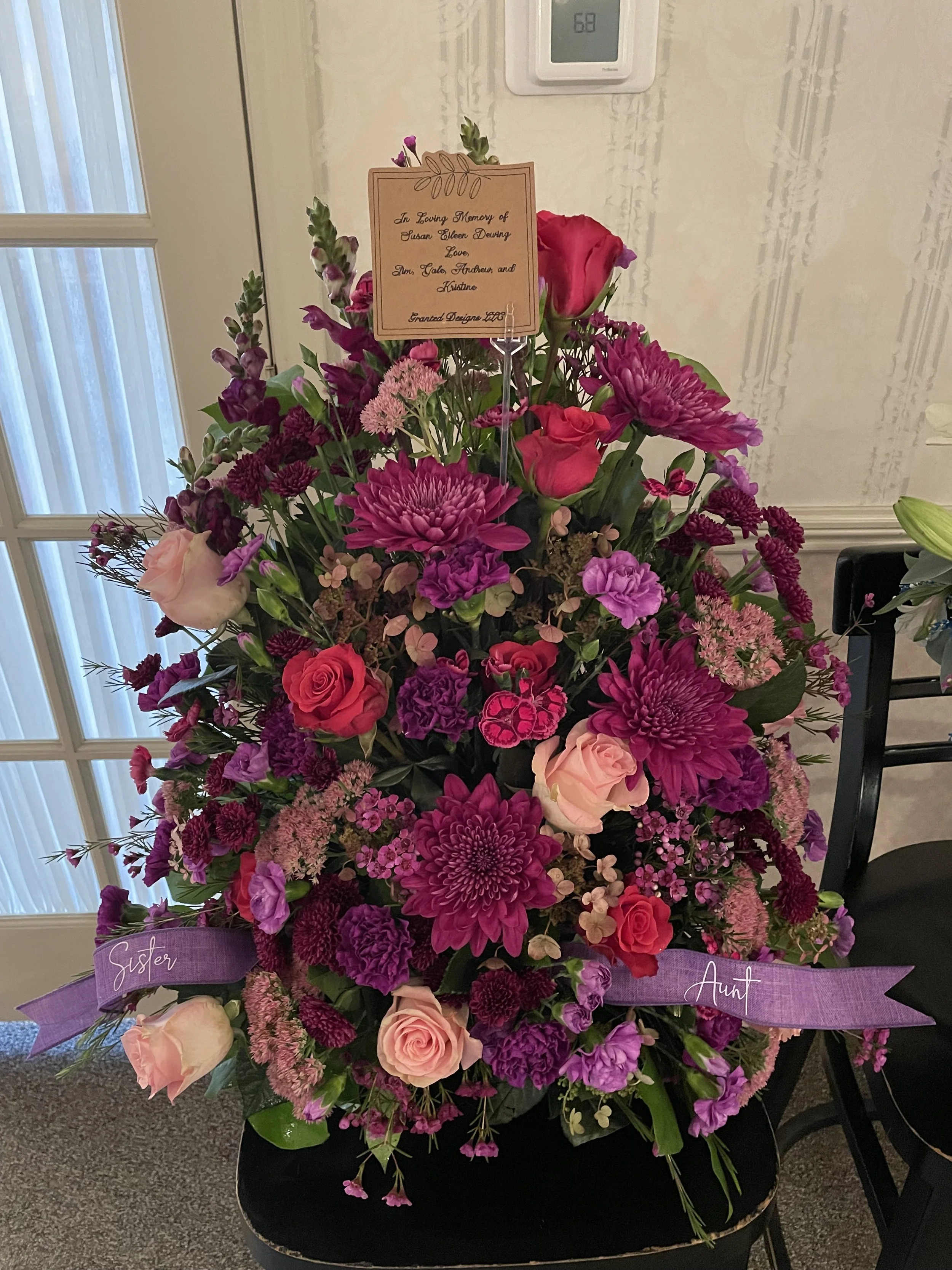A large floral arrangement with pink and purple flowers including roses, chrysanthemums, and carnations, with a sign on top and purple ribbons labeled 'Sister' and 'Aunt'.