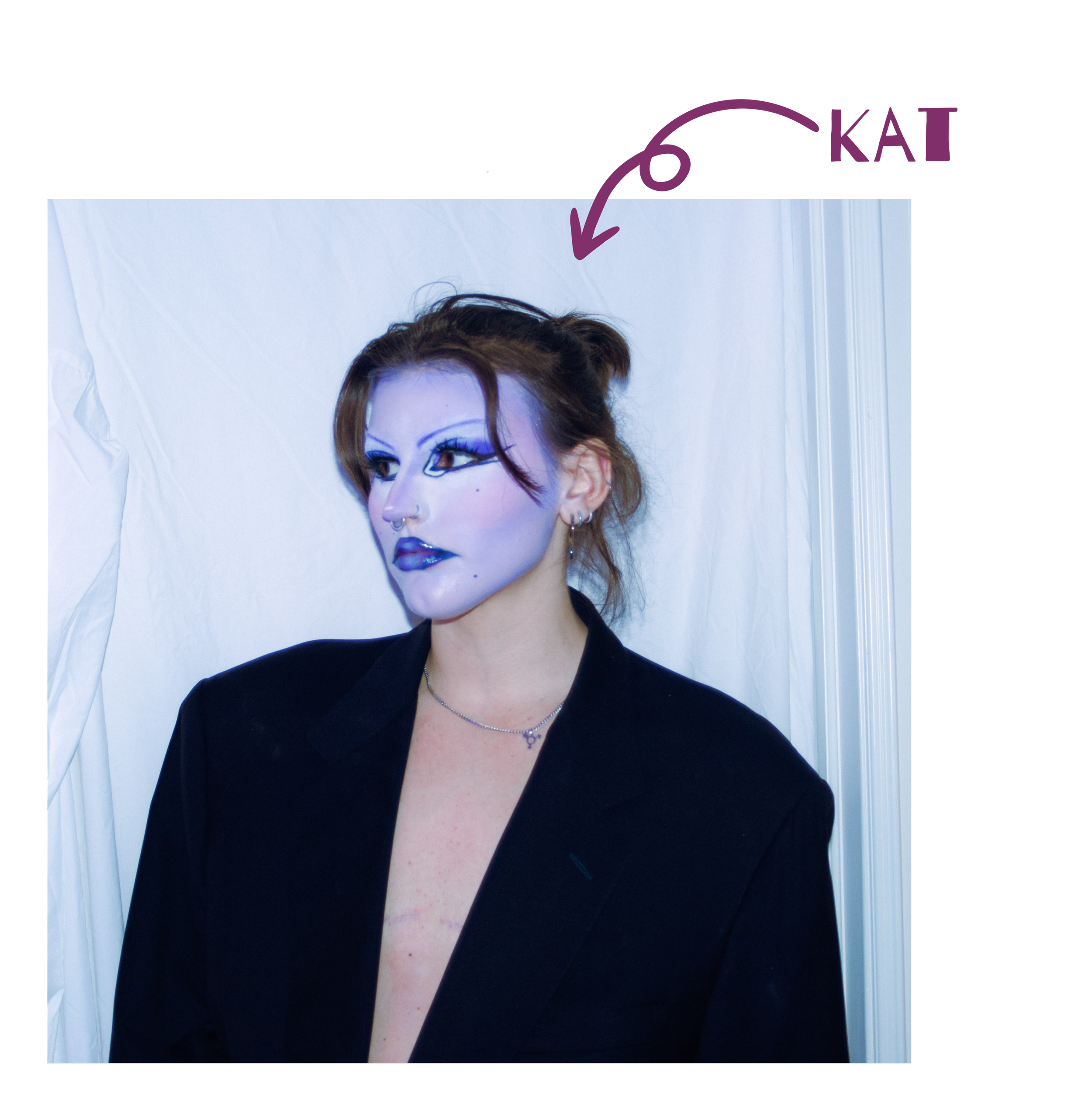 Kat, a white person with brown hair in a bun, stands in front of a white sheet and looks to the side. They wear an oversized blazer over their bare chest, with visible top surgery scars and bold purple and blue makeup.