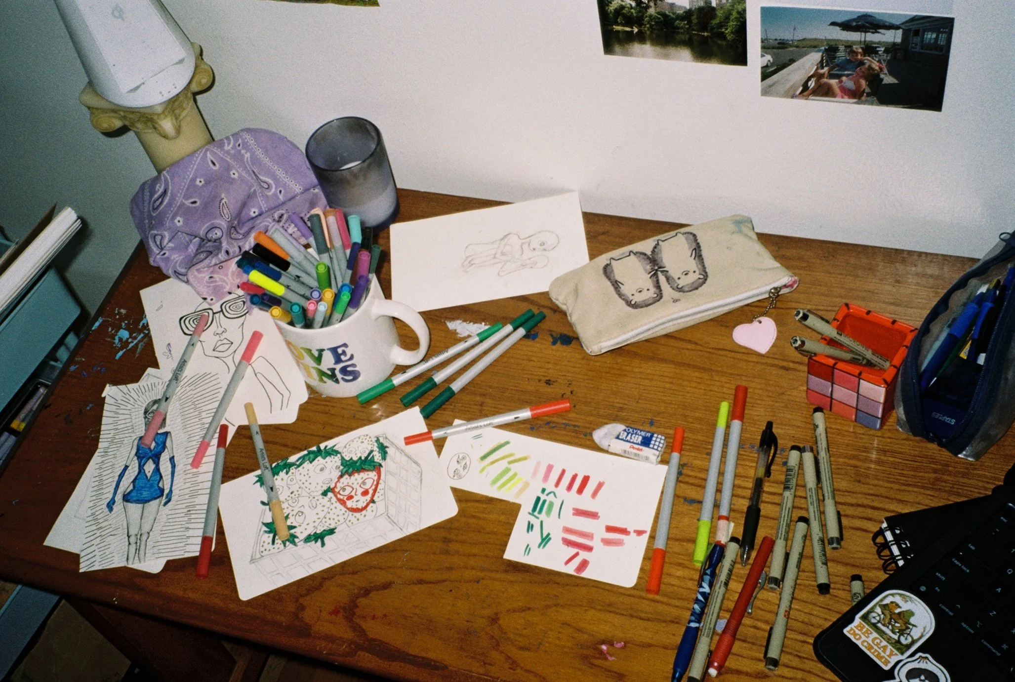 Film photo of Kat’s dark wooden desk covered with markers, pens, and pencils. Half-finished drawings, a pencil case, a candle, and a folded purple bandana are scattered across the surface, showing an active workspace.