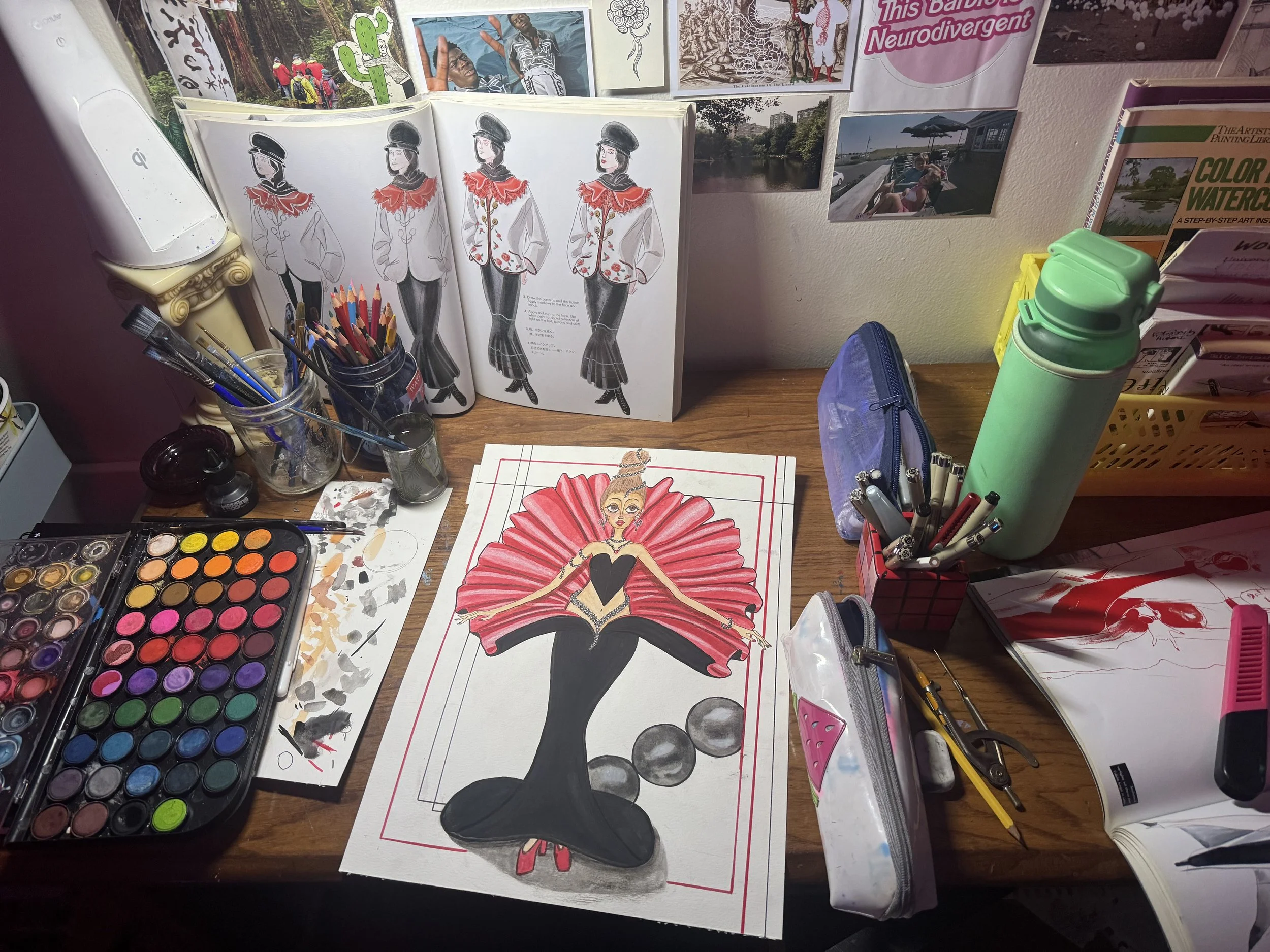 Kat’s desk as they work on a painting centered in the frame, depicting a woman in a black and red corset and gown that fans out behind her tall wig. Surrounding the painting are watercolors, pencils, pens, and an open art book, with small drawings an