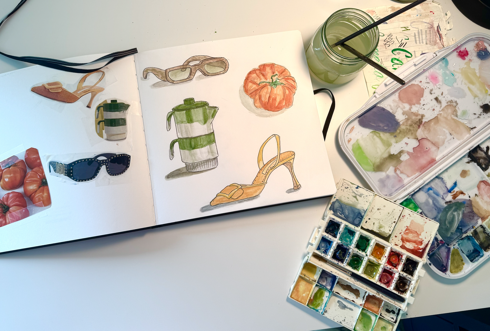 Watercolor palette, paint water, and an open sketchbook viewed from above. On the left page are magazine images taped down. On the right are watercolor studies translating those images, including a pitcher, a high heel, sunglasses, and a tomato.