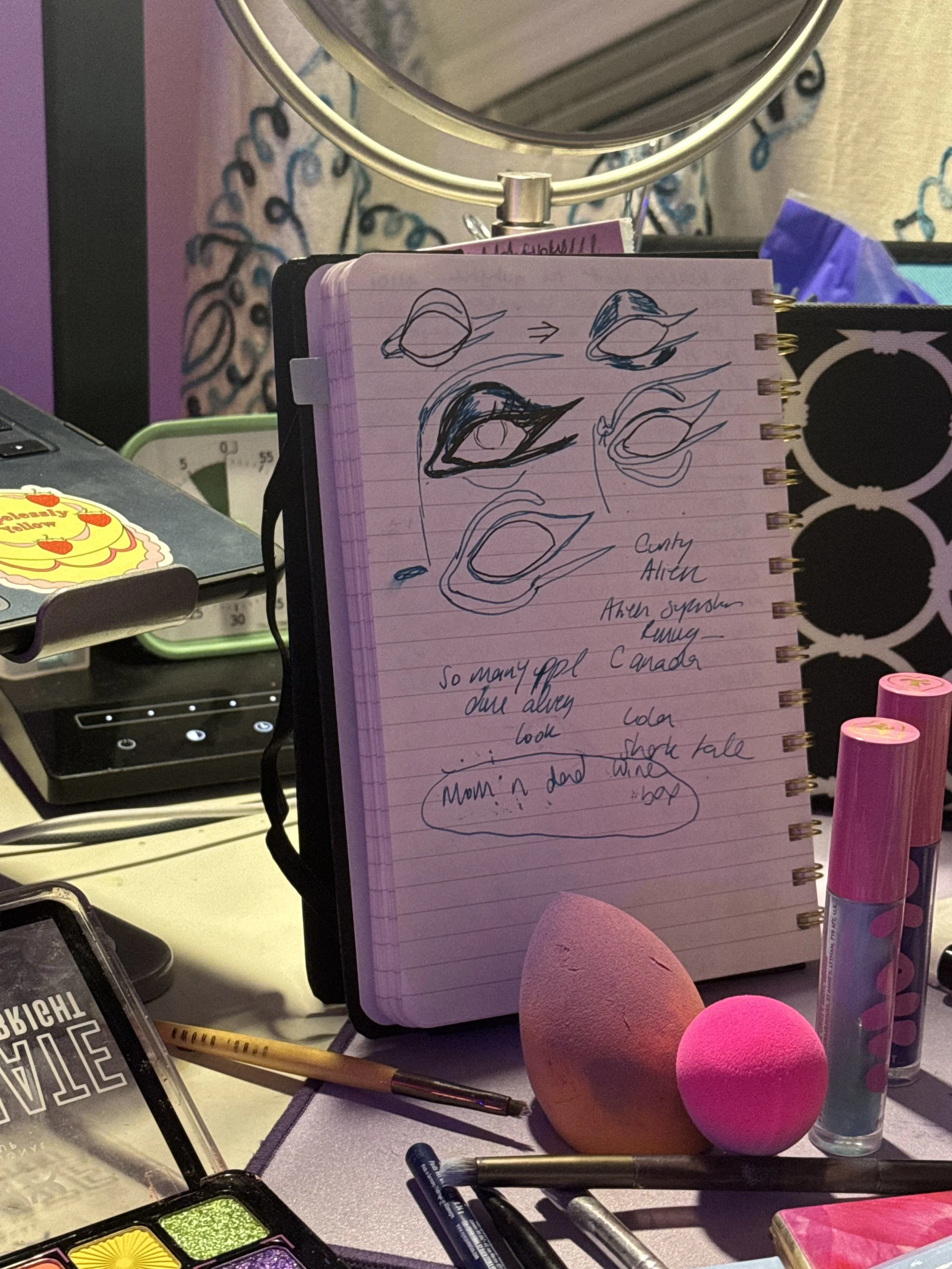 An open notebook filled with sketches exploring an eye shape for drag makeup. Behind the notebook, a mirror and computer sit slightly out of frame. In front are makeup sponges and loose pigments used for experimentation.