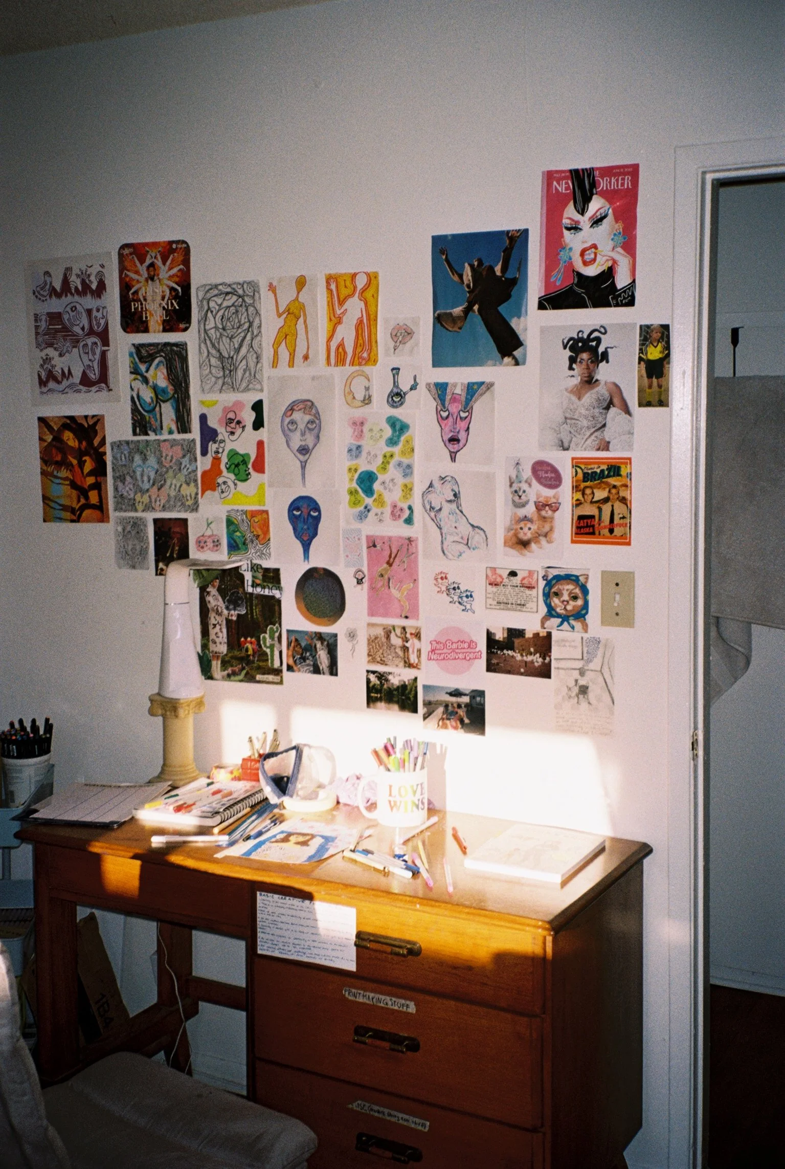 Film photo of Kat’s desk lit by window light, with the wall behind it covered in art, collages, and stickers. Markers, pencils, and notepads are scattered across the desk.