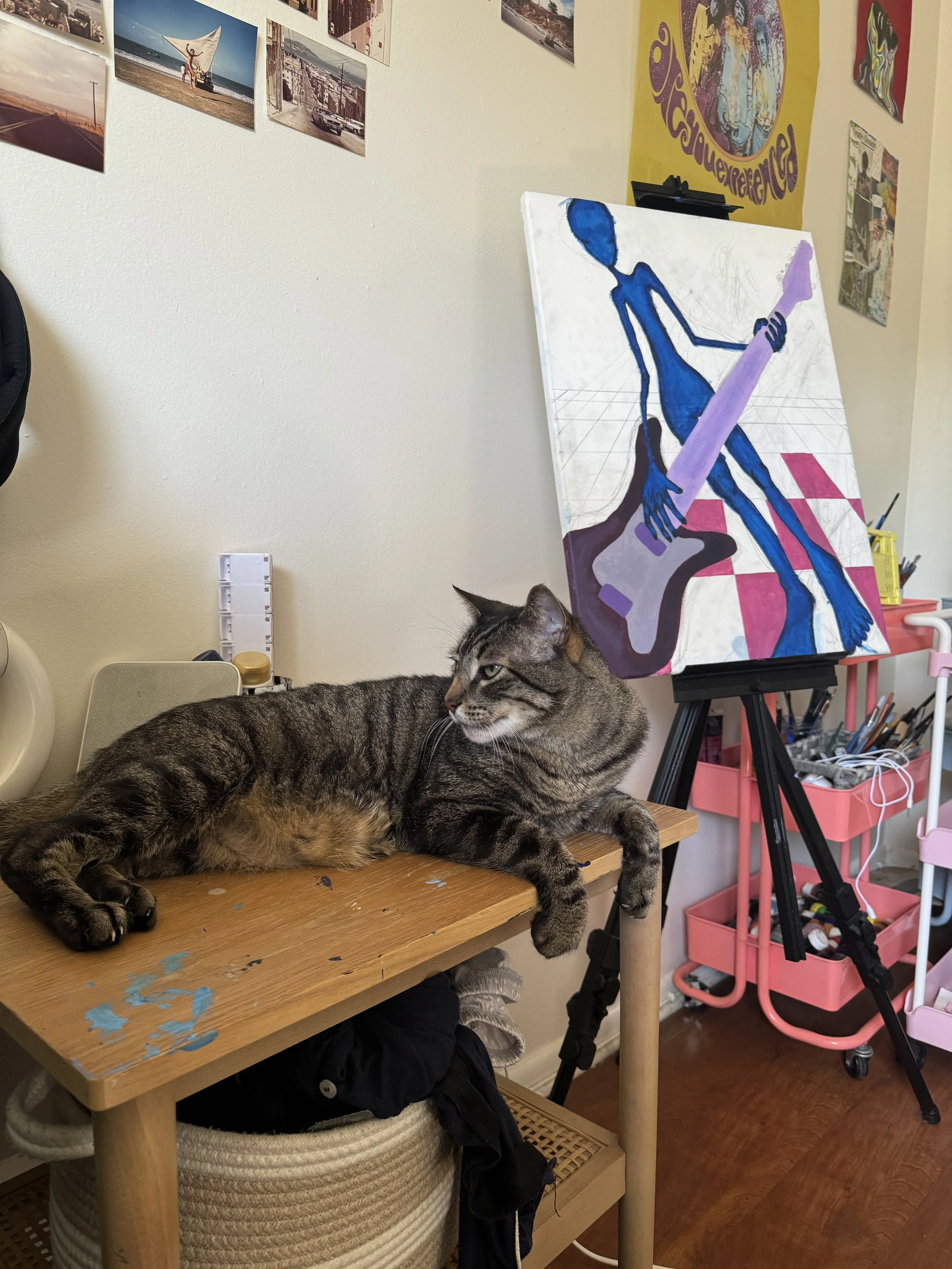 Mike, a striped cat, lies stretched sideways on a small wooden table. Behind him is an easel holding a in-progress painting on canvas and a pink rolling cart filled with art supplies, suggesting a studio space in use.