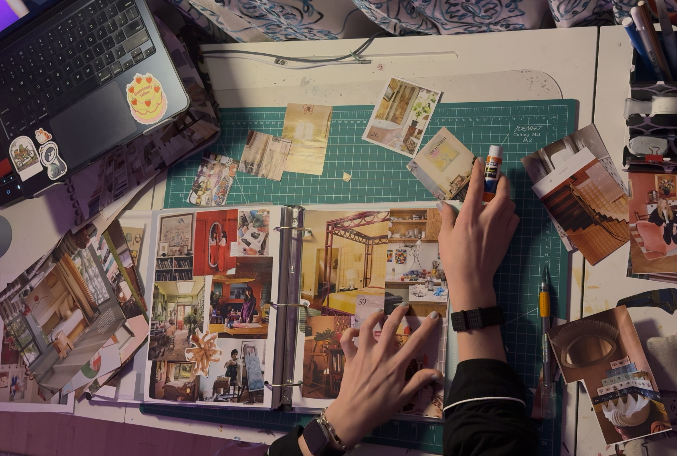 Process photo from above of Kat’s hands collaging on a white desk. A green cutting mat sits beneath a binder filled with glued interior design clippings. Kat holds a glue stick, while an X-Acto knife rests nearby.