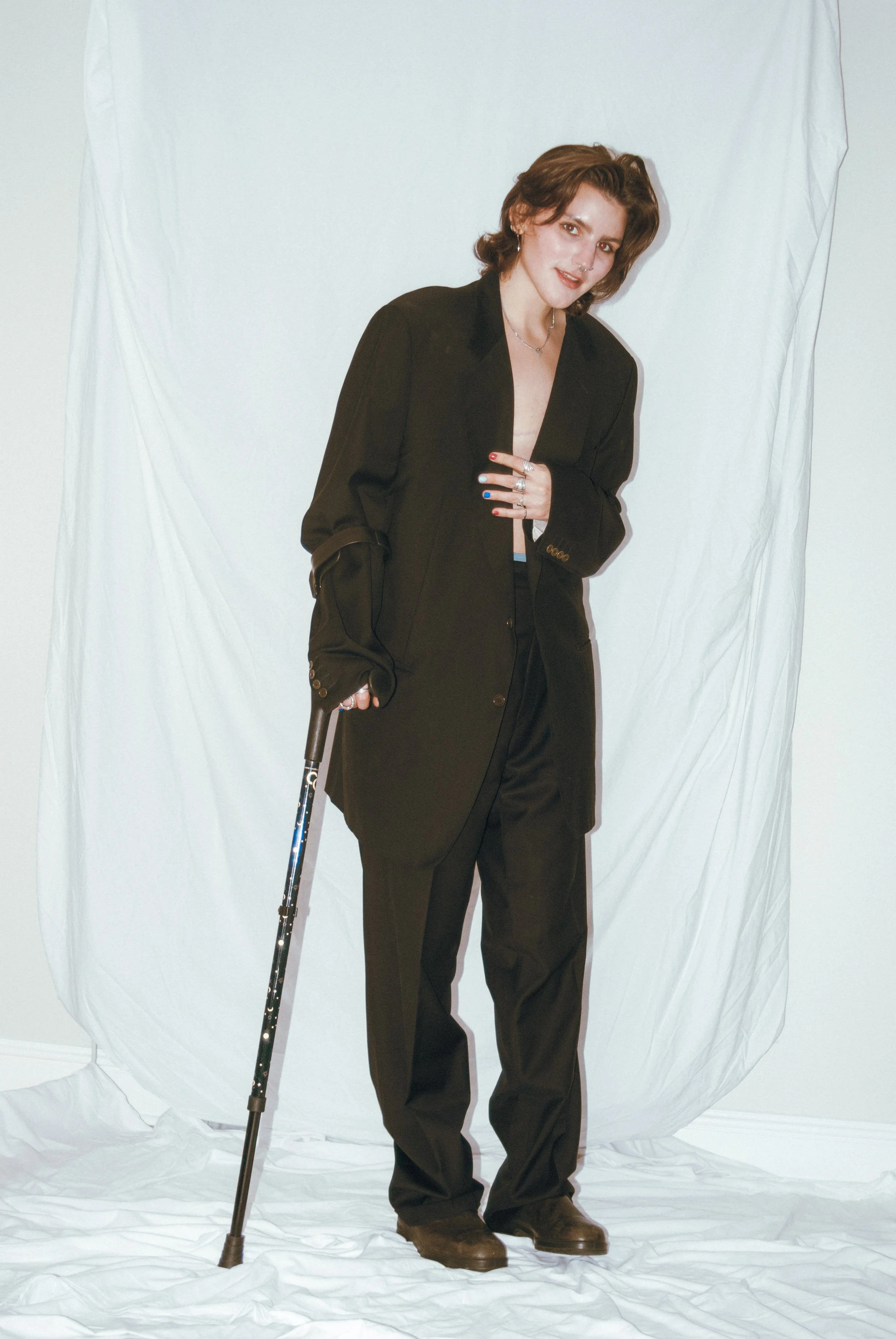 Kat stands in front of a white sheet, slightly smiling and looking toward the camera. They are a white person with short brown hair, wearing an oversized black suit and boots, holding their blazer with one hand and a forearm crutch with the other.