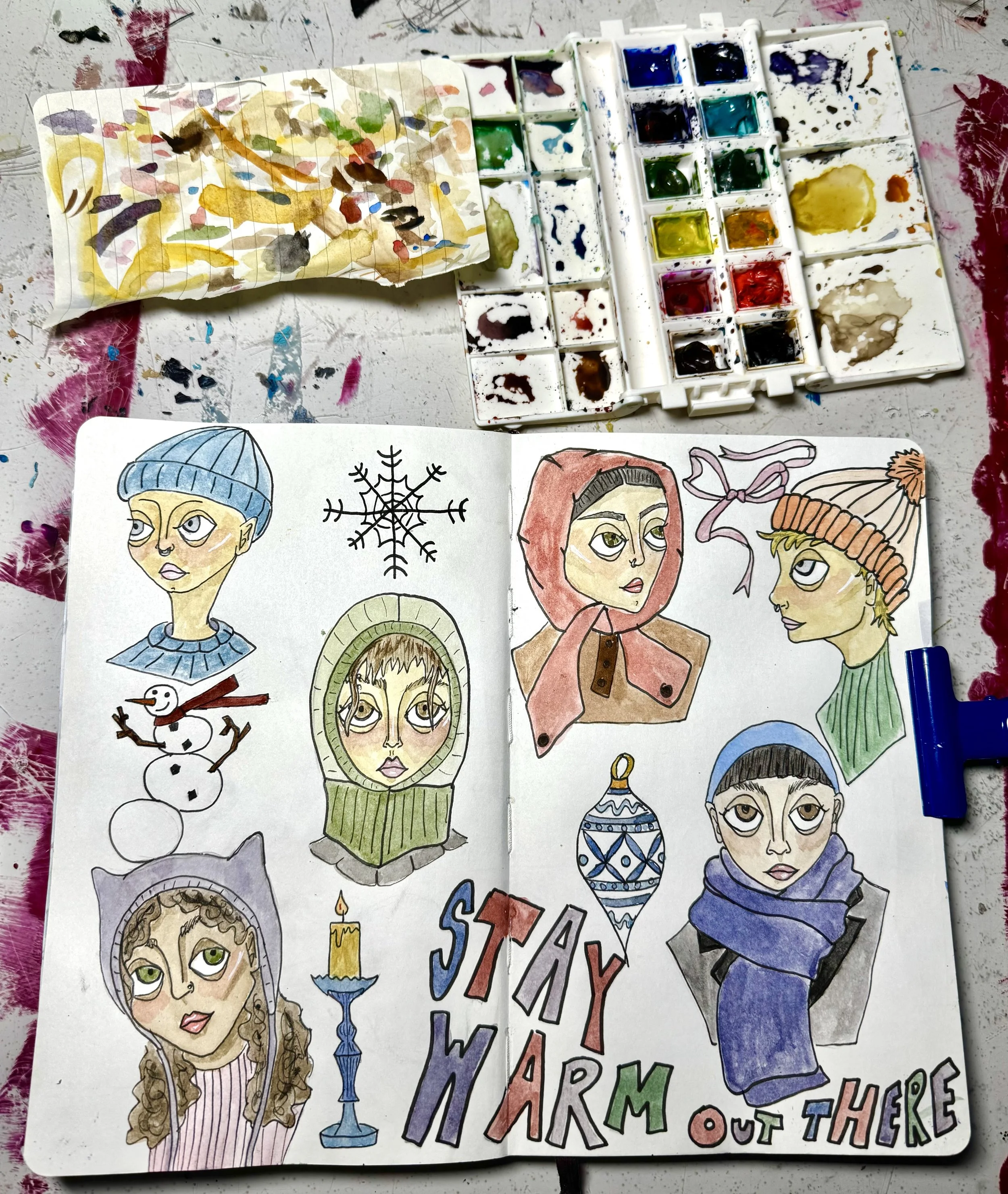 An open sketchbook and watercolor palette viewed from above show six painted busts of people dressed in winter clothing. Hats, scarves, and balaclavas appear in blues, greens, reds, and purples. Between the figures are winter motifs like ribbons, orn