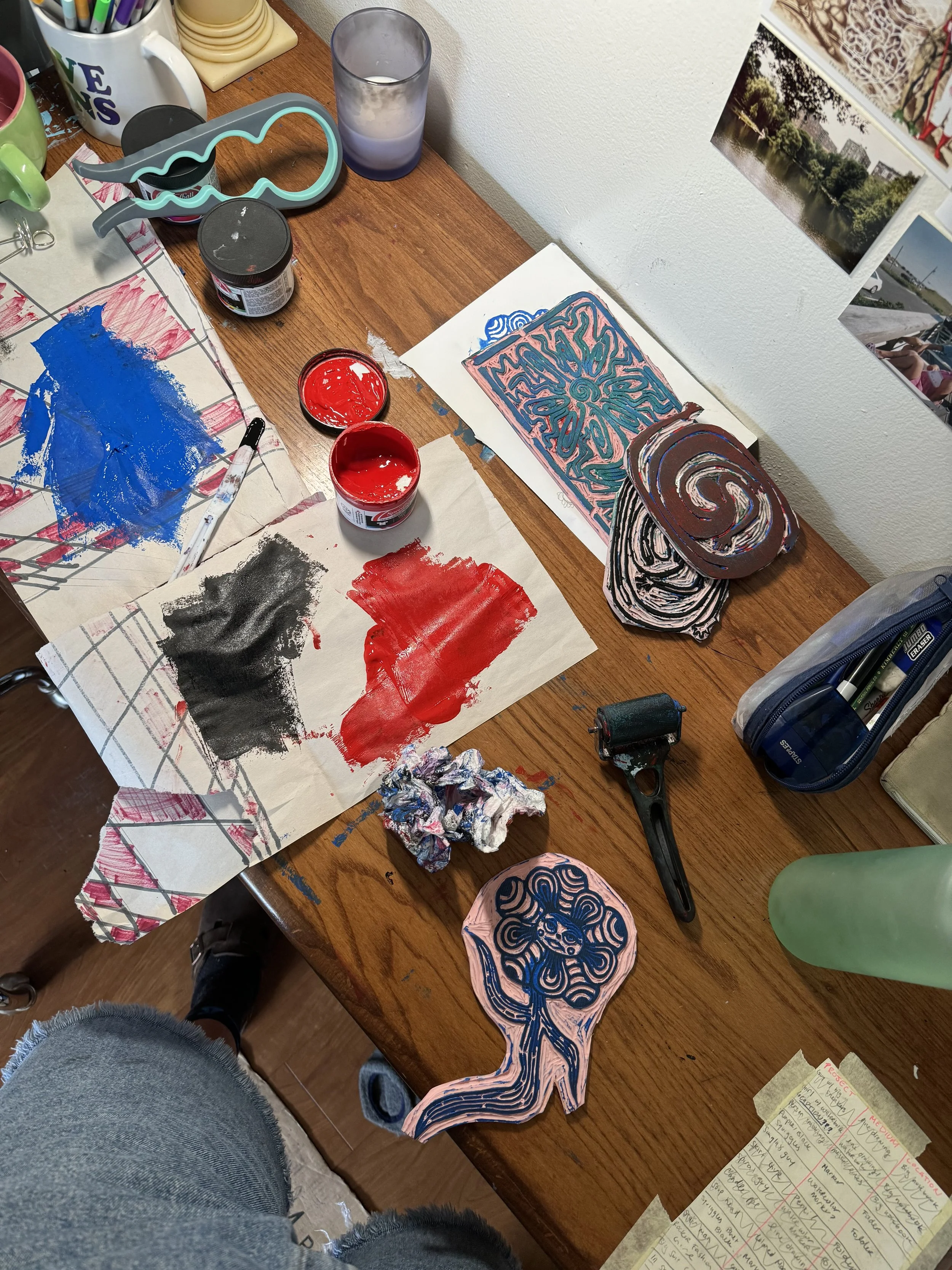 Kat’s desk viewed from above as they work on linocut prints. The dark wooden surface holds carved linoleum stamps, ink, an ink roller, a candle, and scrap paper smeared with black ink.