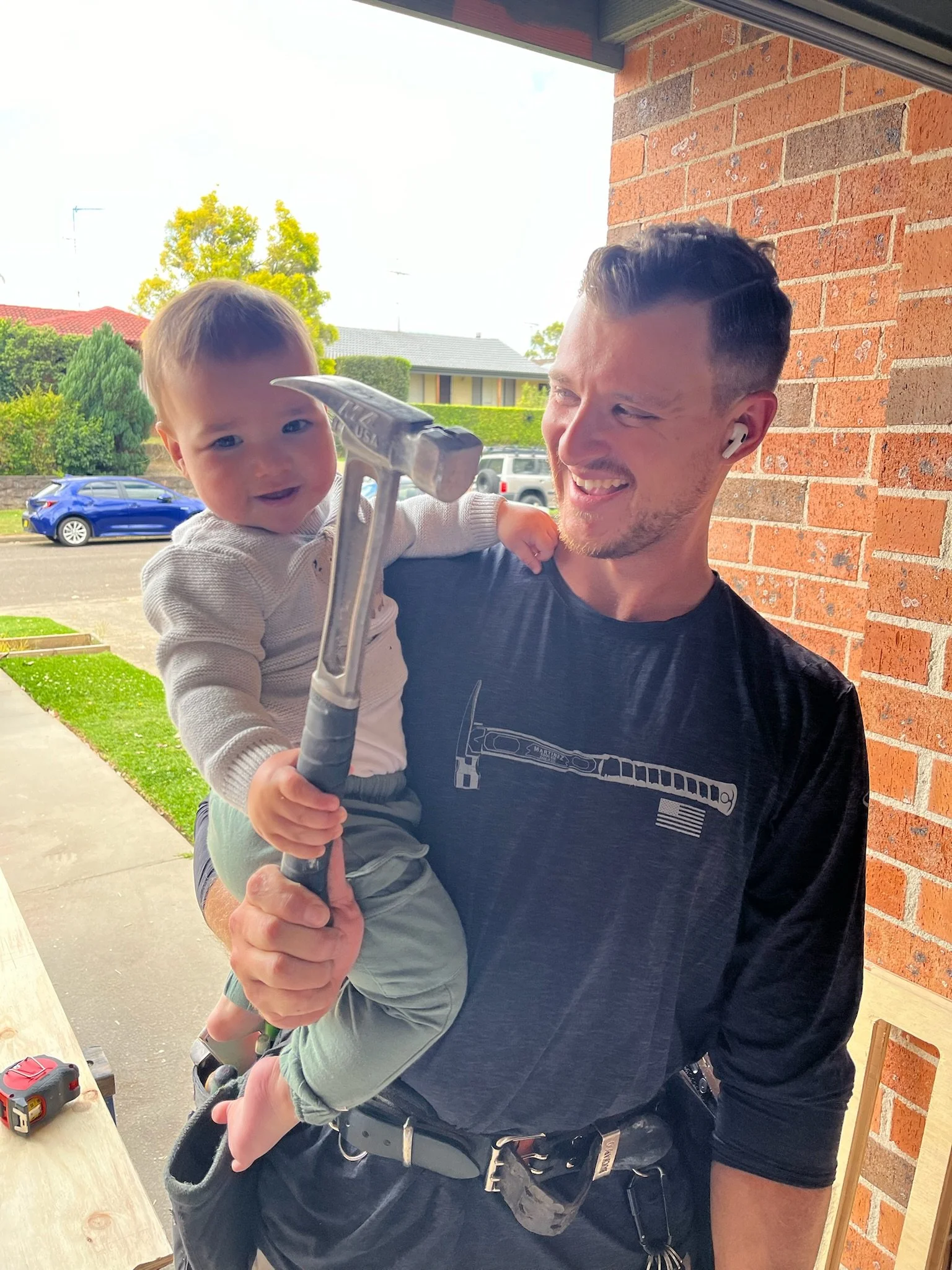 A man holding a smiling toddler with a hammer inside a house, with a car and houses outside in the background.