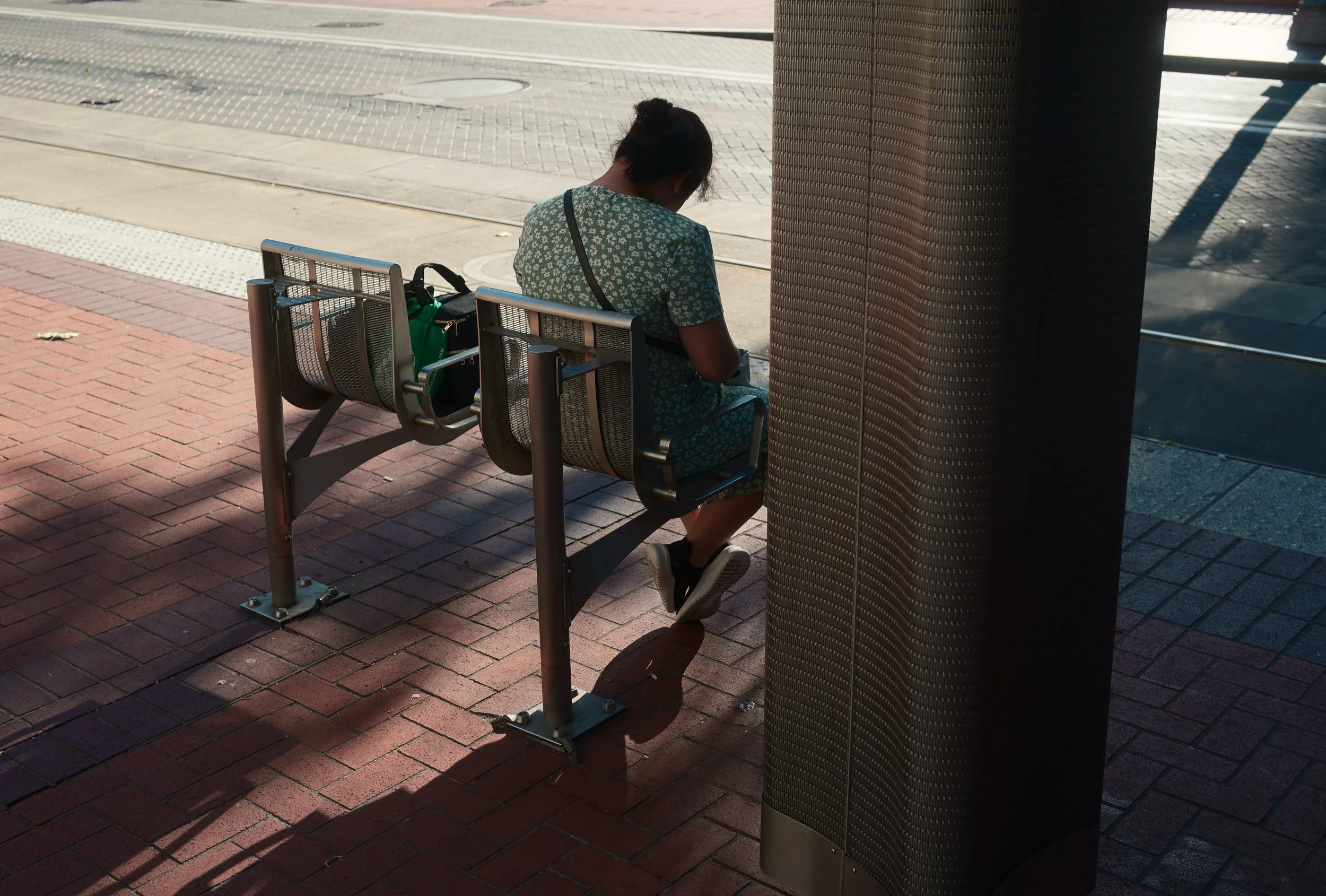 A young girl with dark hair tied in a bun, wearing a green and white floral dress, sitting on a metal bench at a bus stop or outdoor seating area on a sunny day. She appears to be looking at her phone or a similar device. The background shows a sidew