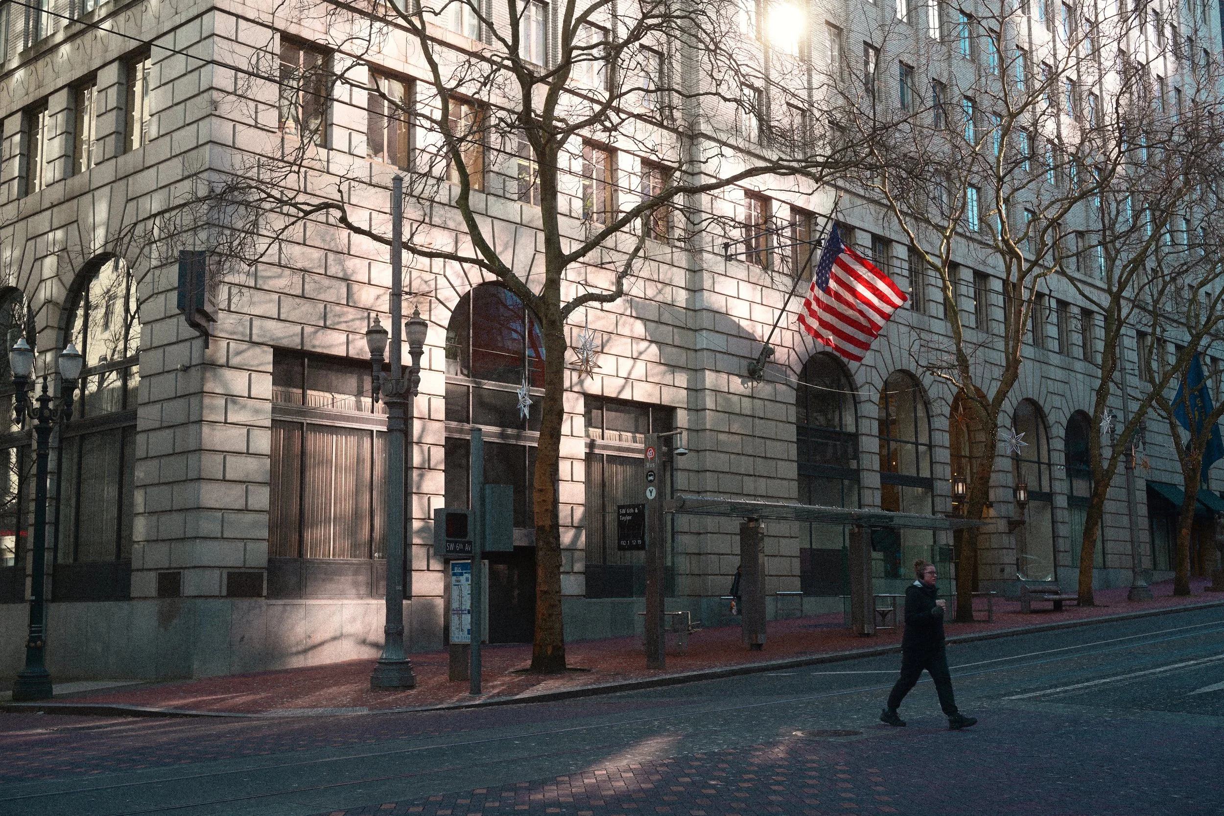 Downtown Portland street with an American flag, historic stone buildings, leafless trees, bus stop, and pedestrian.