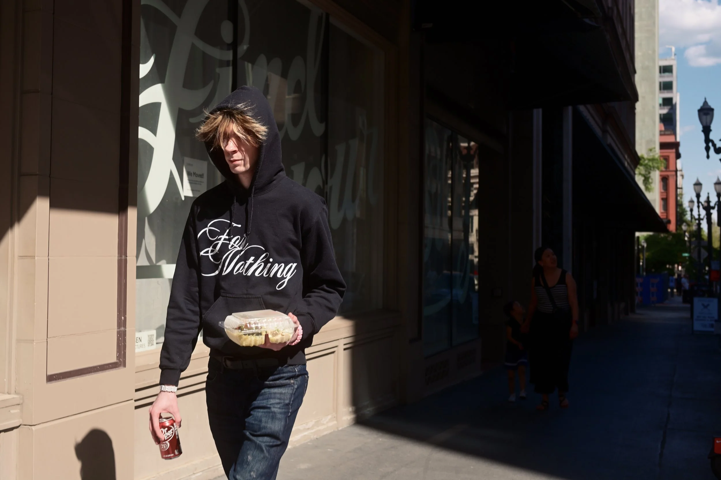 A person wearing a black hoodie with the text "Nothing" on it, holding a red can of Dr. Pepper and a container of food, walking past a storefront on a city street.