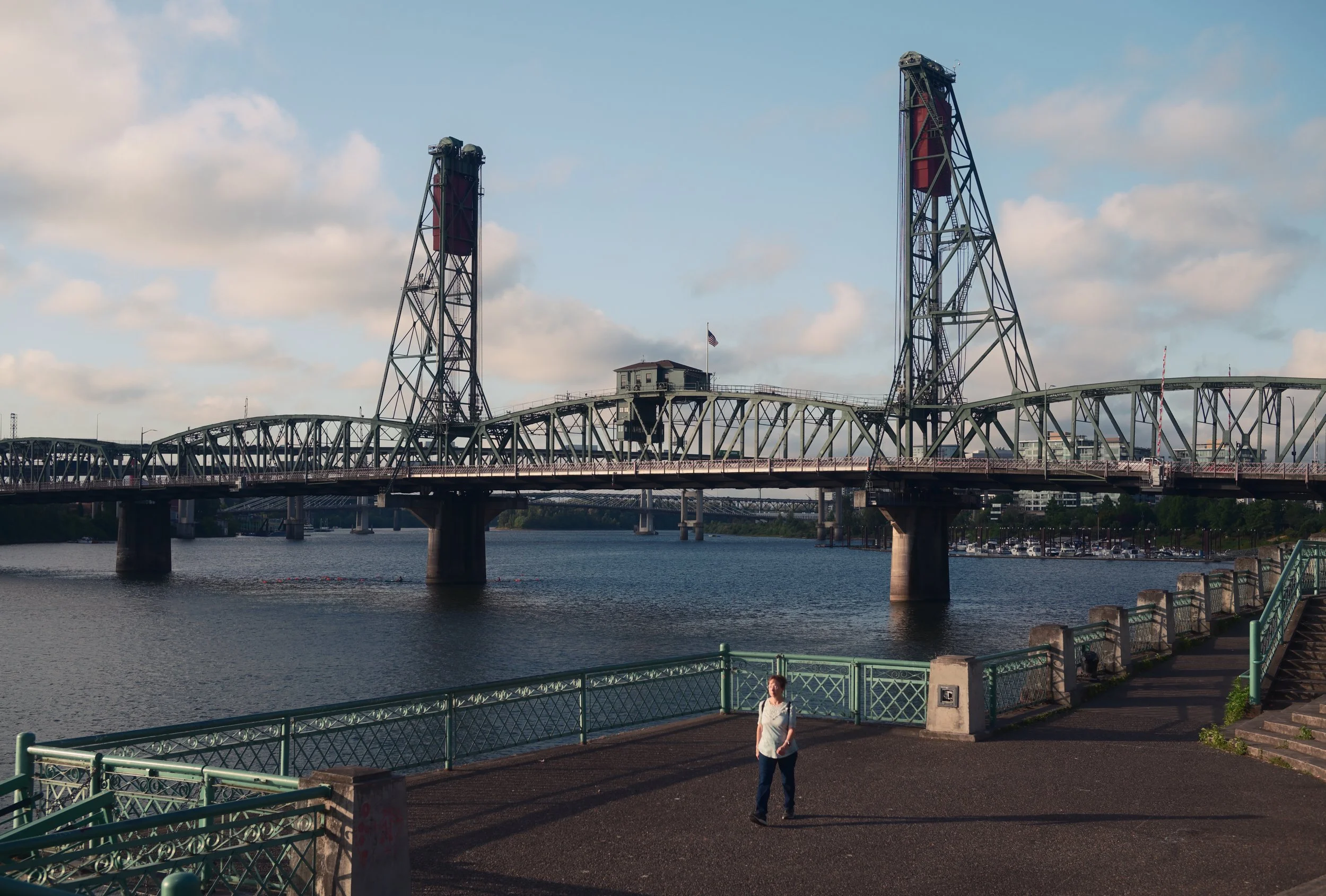A woman standing near the water with a bridge in the background. The bridge has two large towers and a truss structure across the water. The sky is partly cloudy with the sun casting shadows.