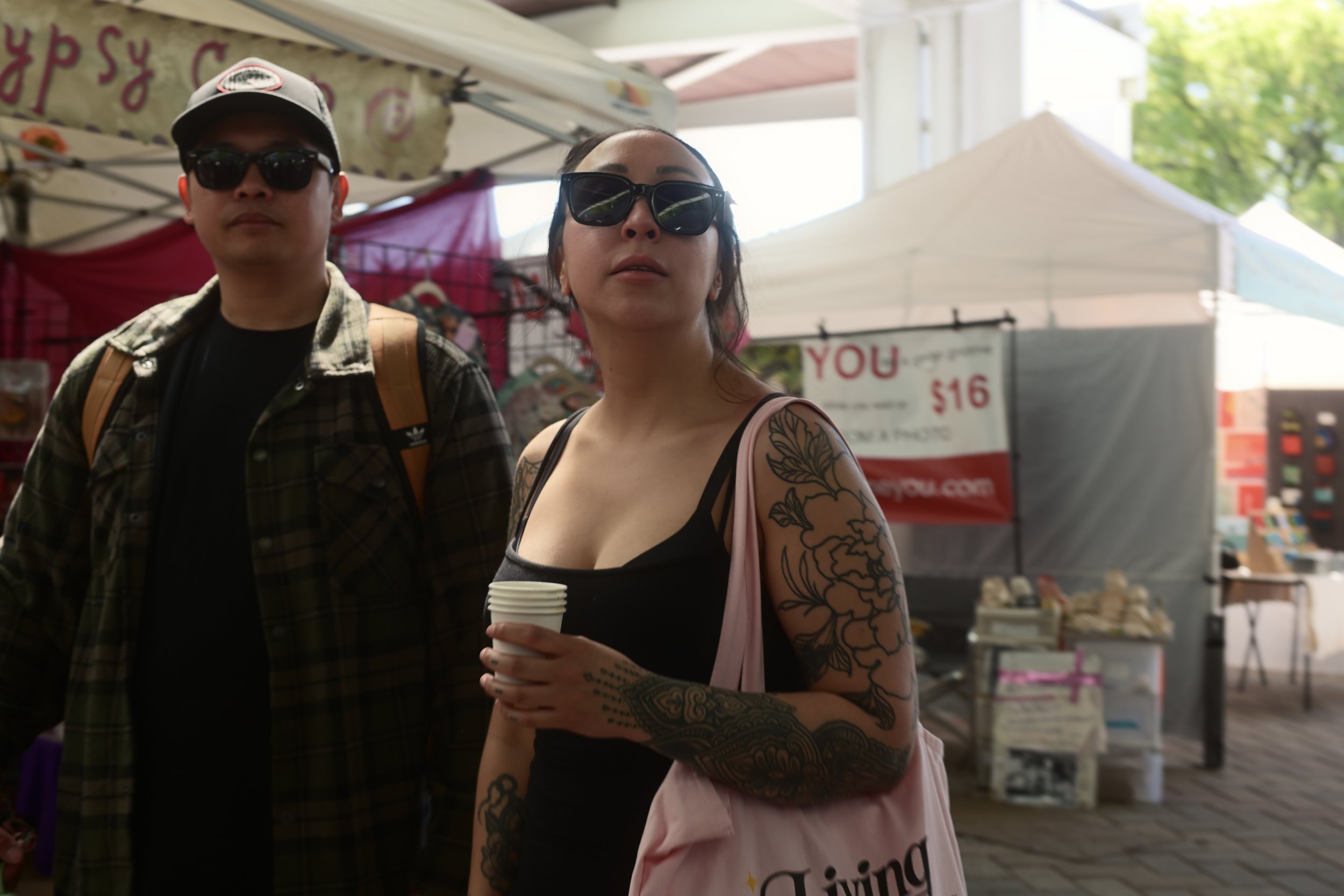 Two people, a man and a woman, standing outdoors at a market or fair. Both wearing sunglasses and facing the camera. The woman has tattoos on her arms and is holding a small coffee cup. The background shows tents and signs, with green trees and sunlight visible.