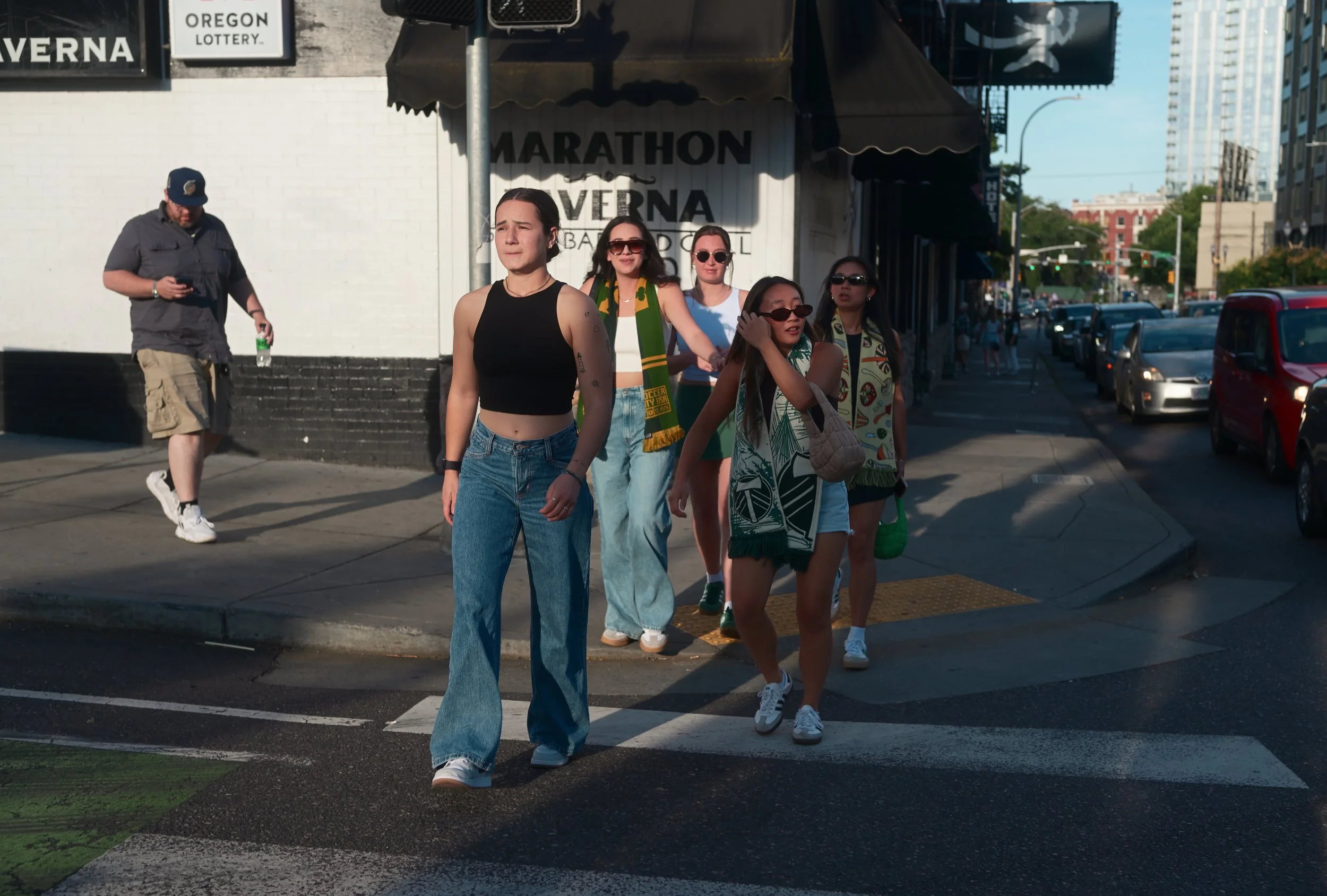 Group of young women walking across a crosswalk on a city street in late afternoon sunlight, with a man in the background looking at his phone, and buildings and cars along the street.