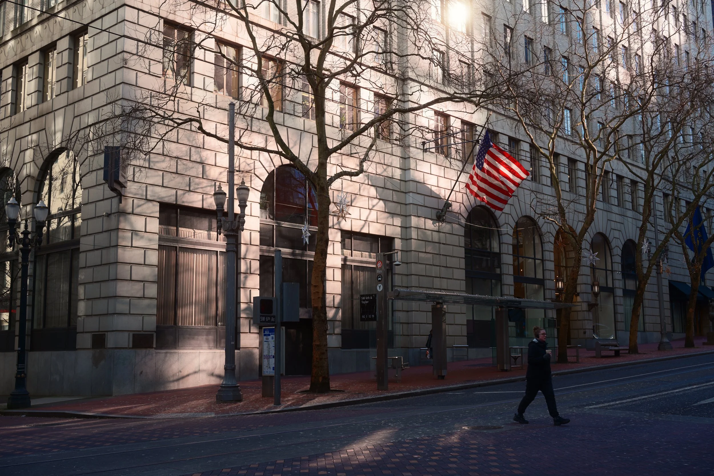 A street view featuring a building with arched windows and an American flag hanging outside. Bare trees line the sidewalk, and a person is walking across the street during daytime.