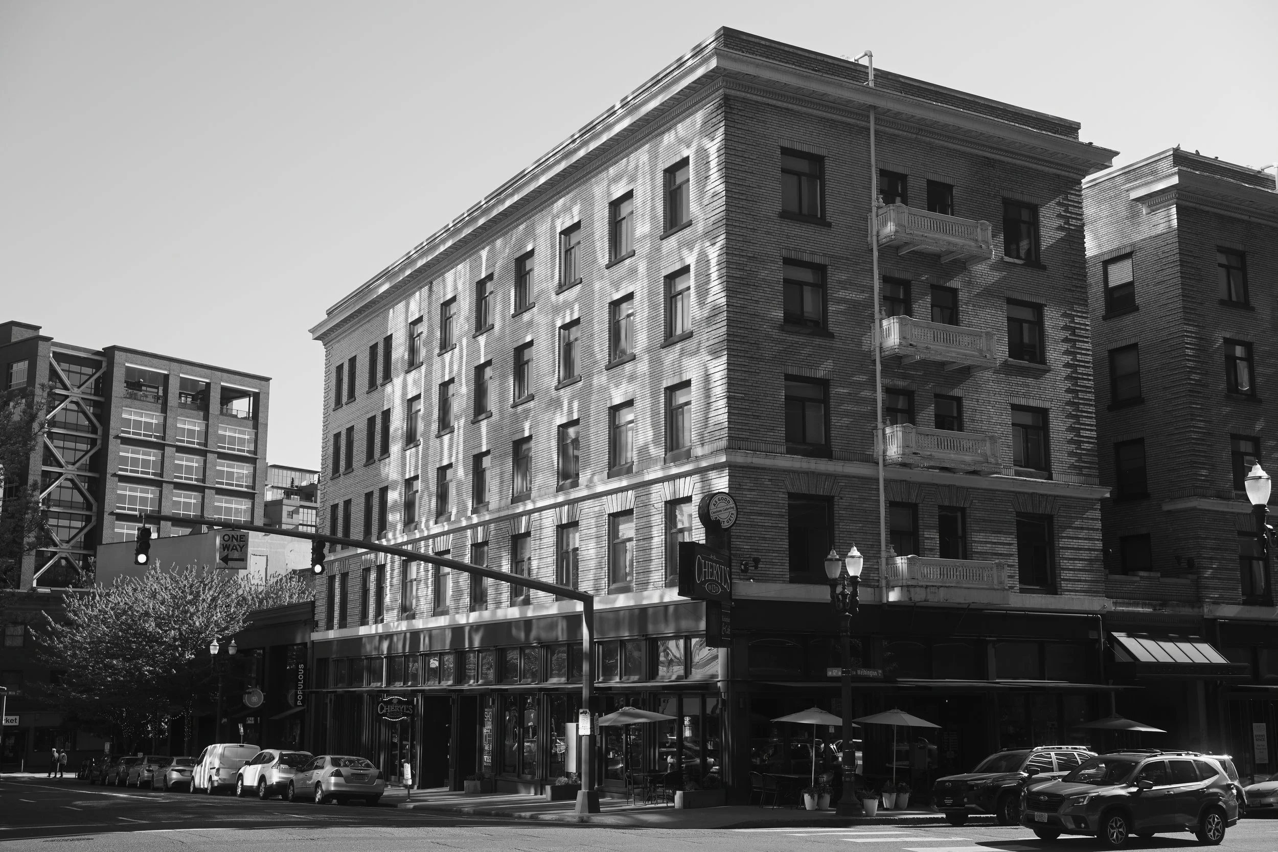 A black and white photo of a multi-story building with brick exterior, featuring balconies on the upper floors, street-level storefronts, and parked cars in front.
