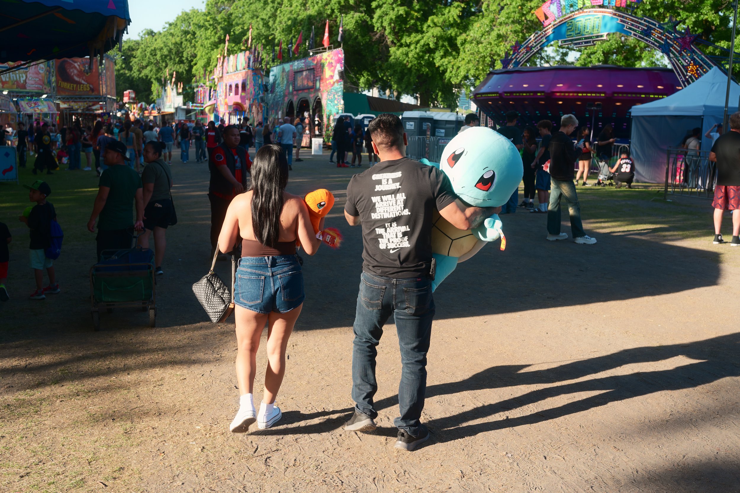 People walking through a carnival with rides and colorful booths, including a funhouse and a spinning ride, on a sunny day surrounded by green trees.