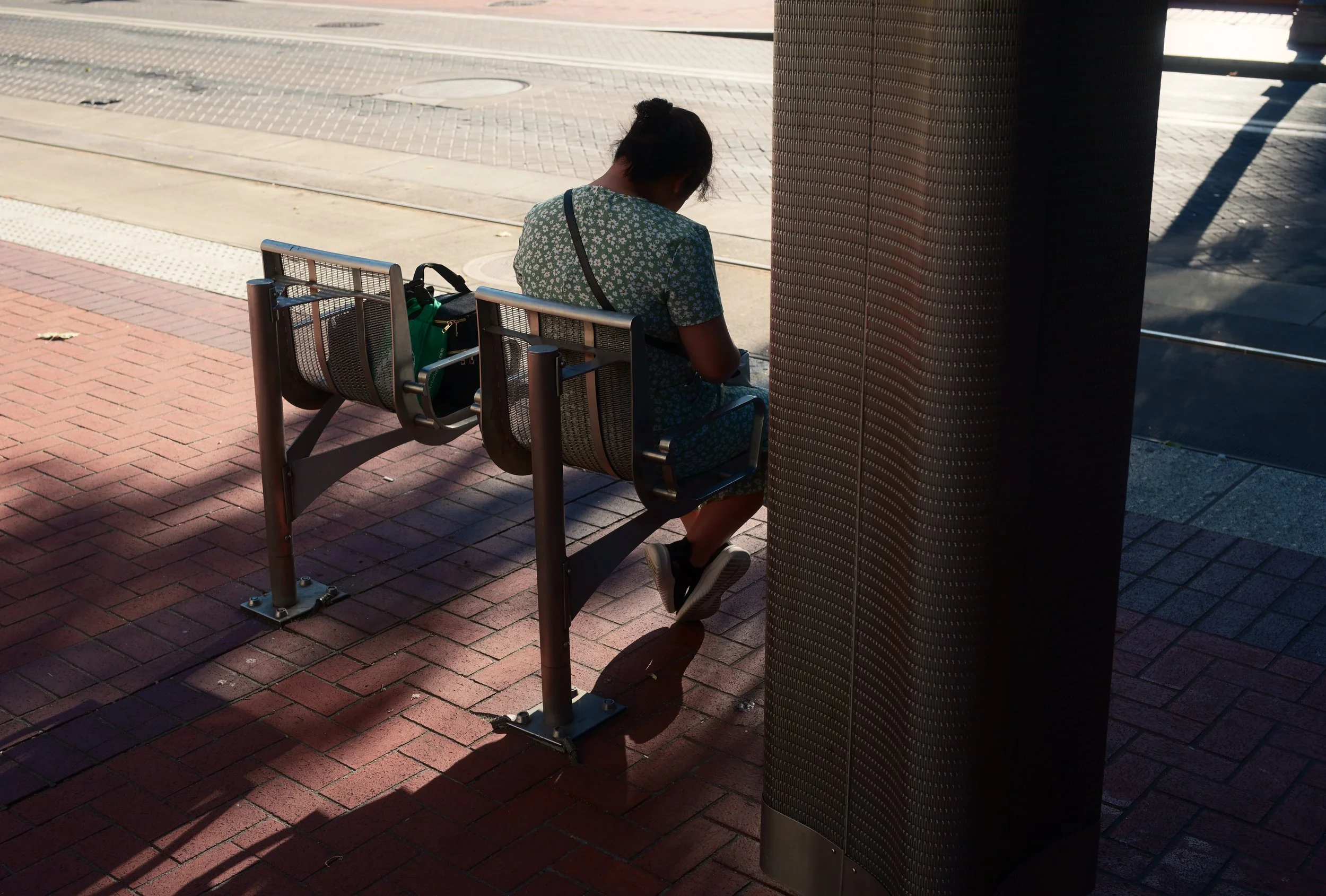 A woman sitting on a bench at a bus stop, looking at her phone. She has dark hair tied up, and she is wearing a floral dress and sneakers. The area has red brick flooring and a large dark column on the right.