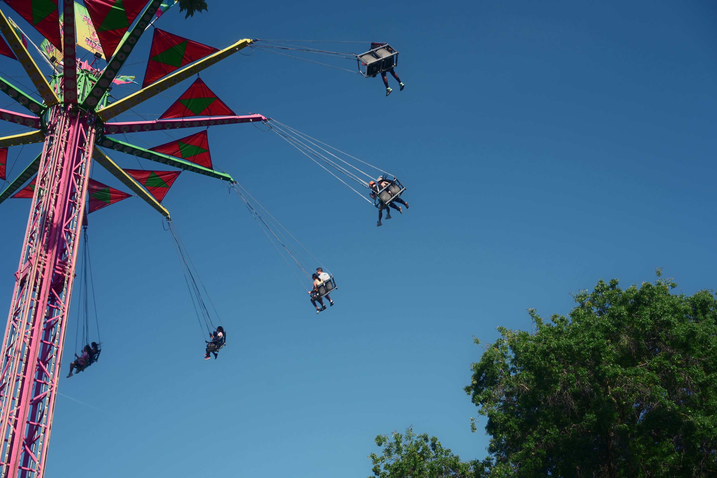 Colorful amusement park swing ride with people sitting in seats, swinging high with blue sky and green trees in the background.