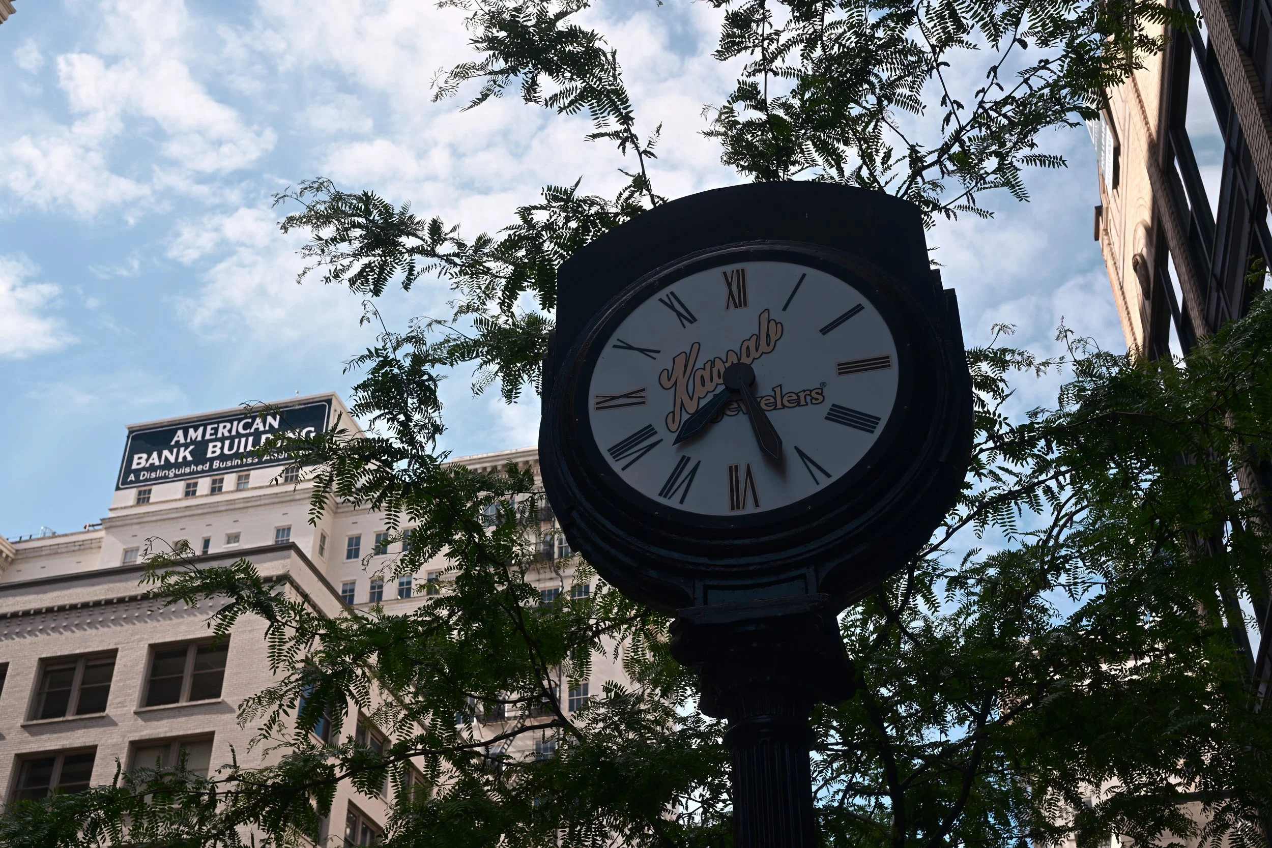 A clock on a street corner displays the time as approximately 4:44. The clock has Roman numerals and is surrounded by trees and buildings on a partly cloudy day.