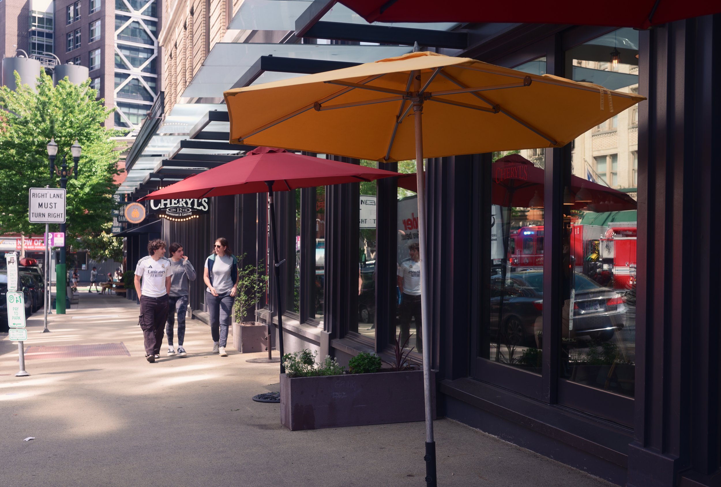 Pedestrians walking past a restaurant with red and yellow umbrellas outside on a sunny day.