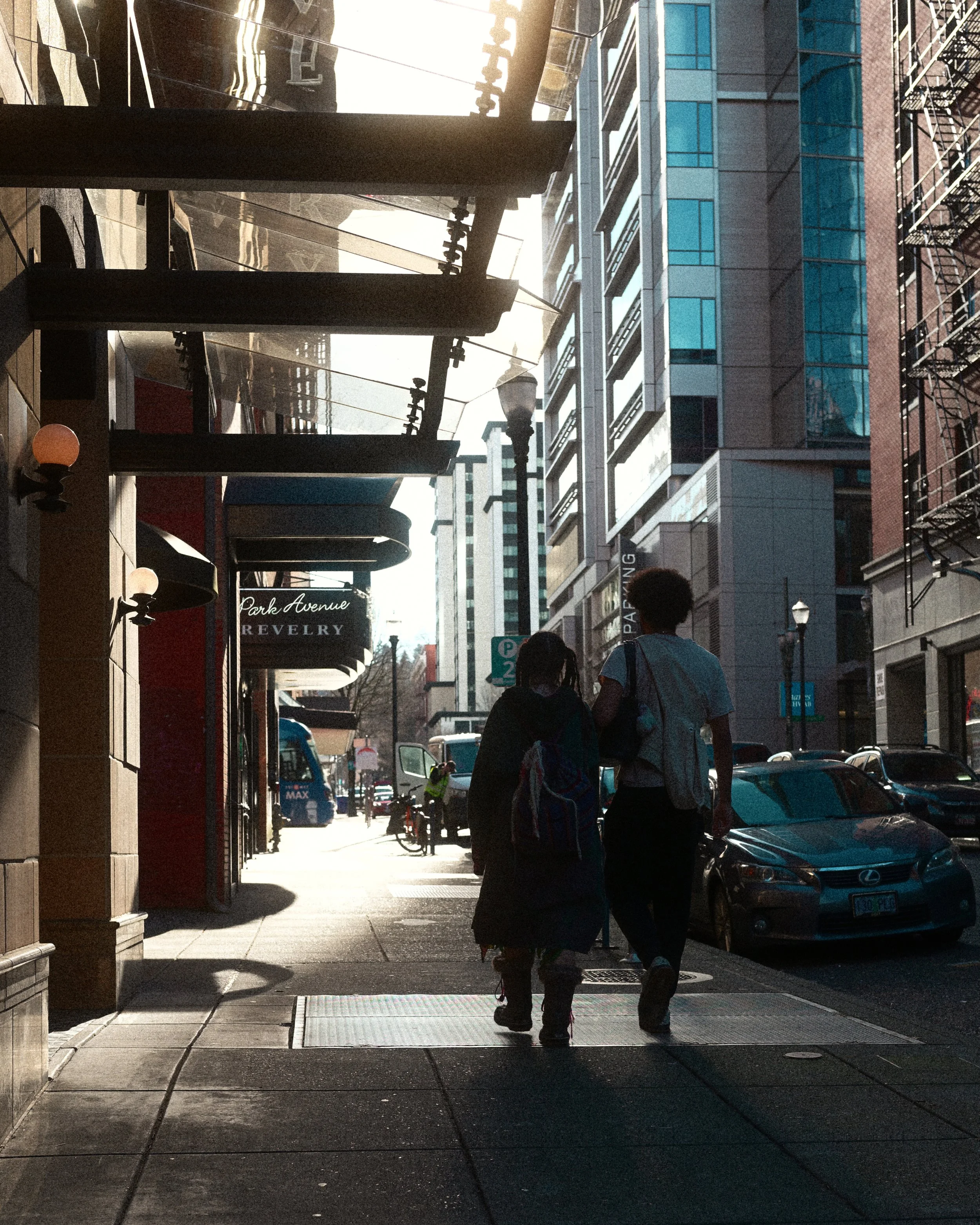 Pedestrians walking through a downtown city street beneath elevated transit tracks, surrounded by high-rise buildings and parked cars.