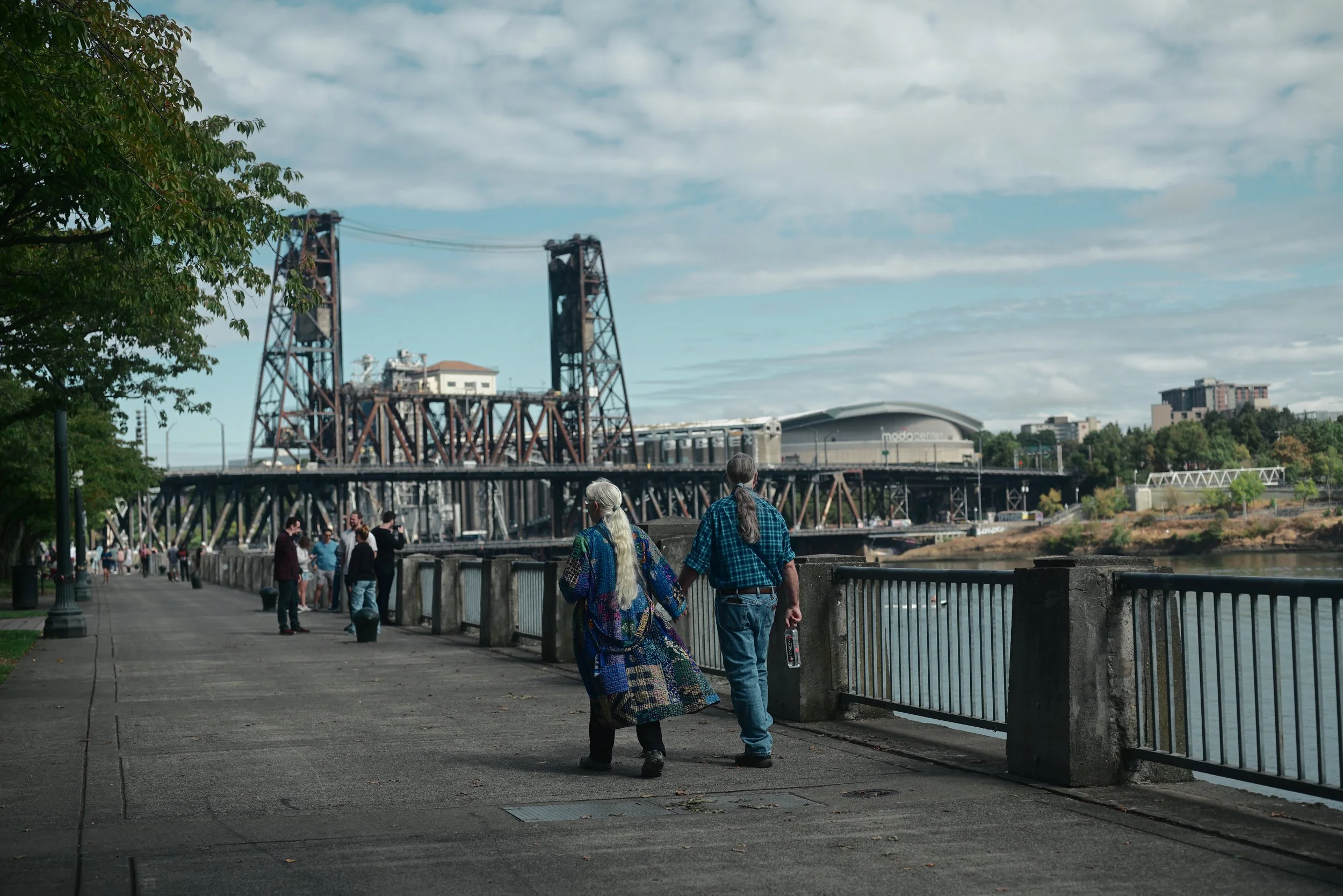 People walking along a riverside promenade with a bridge and city buildings in the background.
