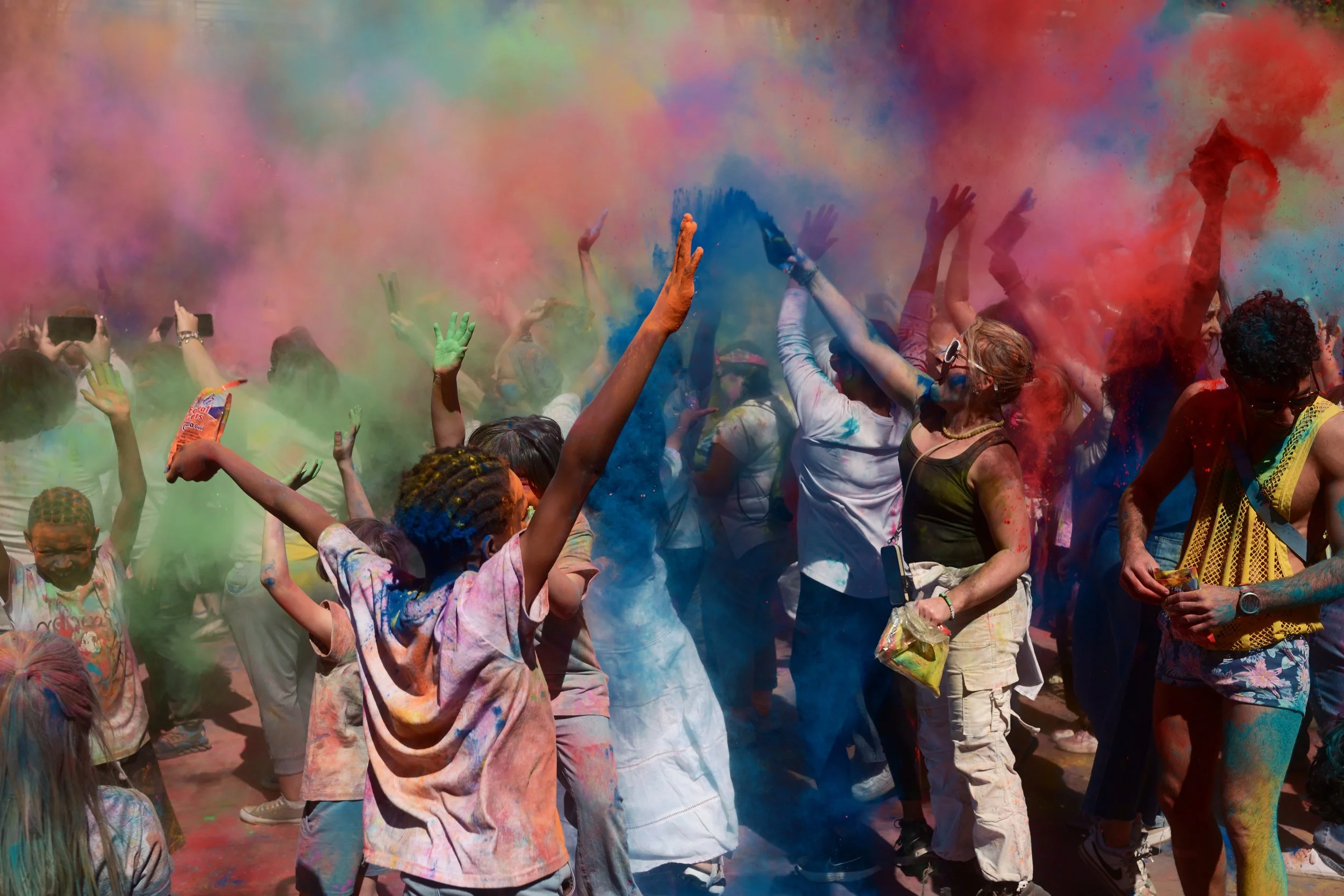 People celebrating during the Holi festival, throwing colorful powder into the air, with vibrant pink, blue, green, and red powders creating a lively scene.