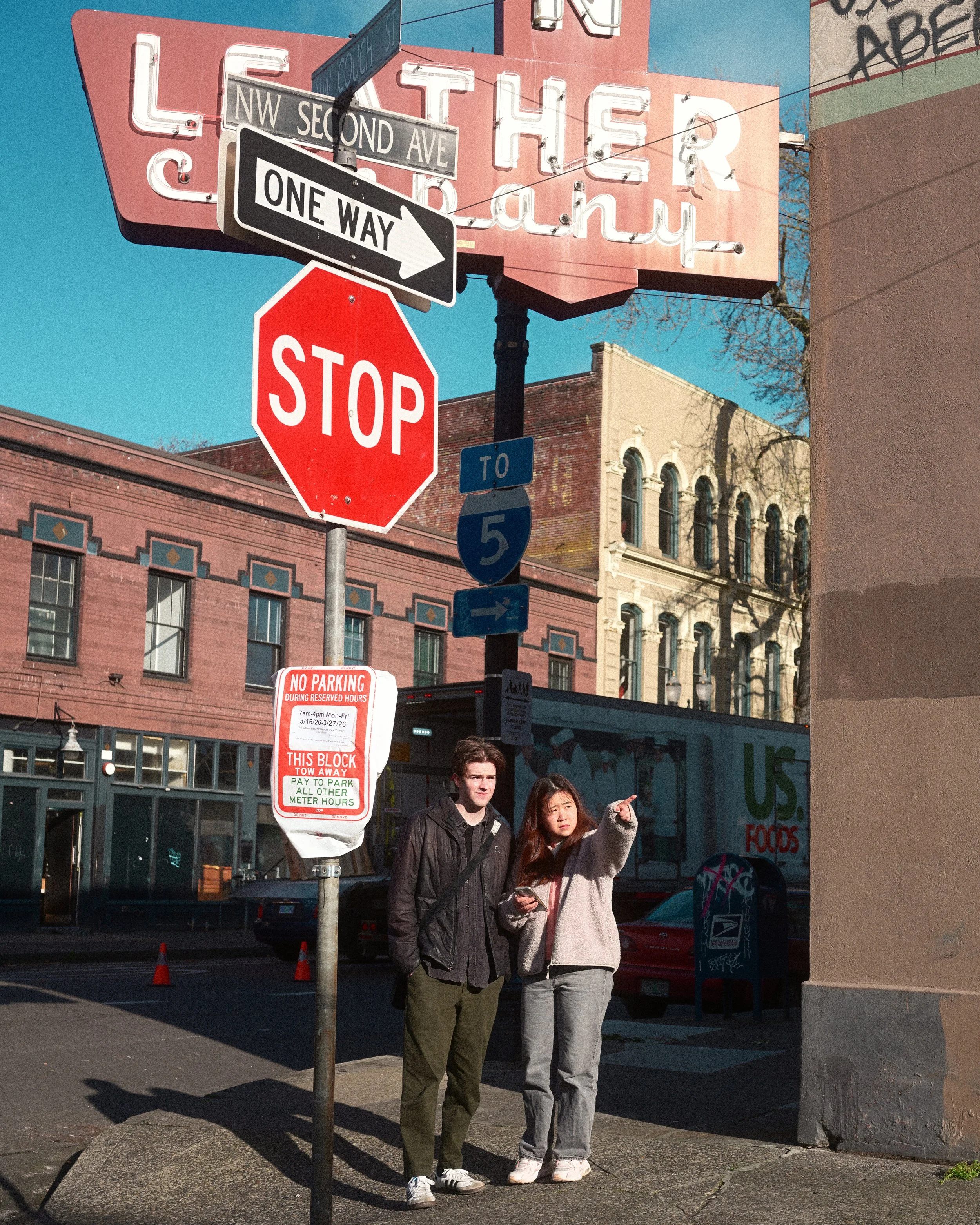 Two pedestrians stand beneath vintage street signs reading NW Second Ave, One Way, and Stop in a downtown district.
