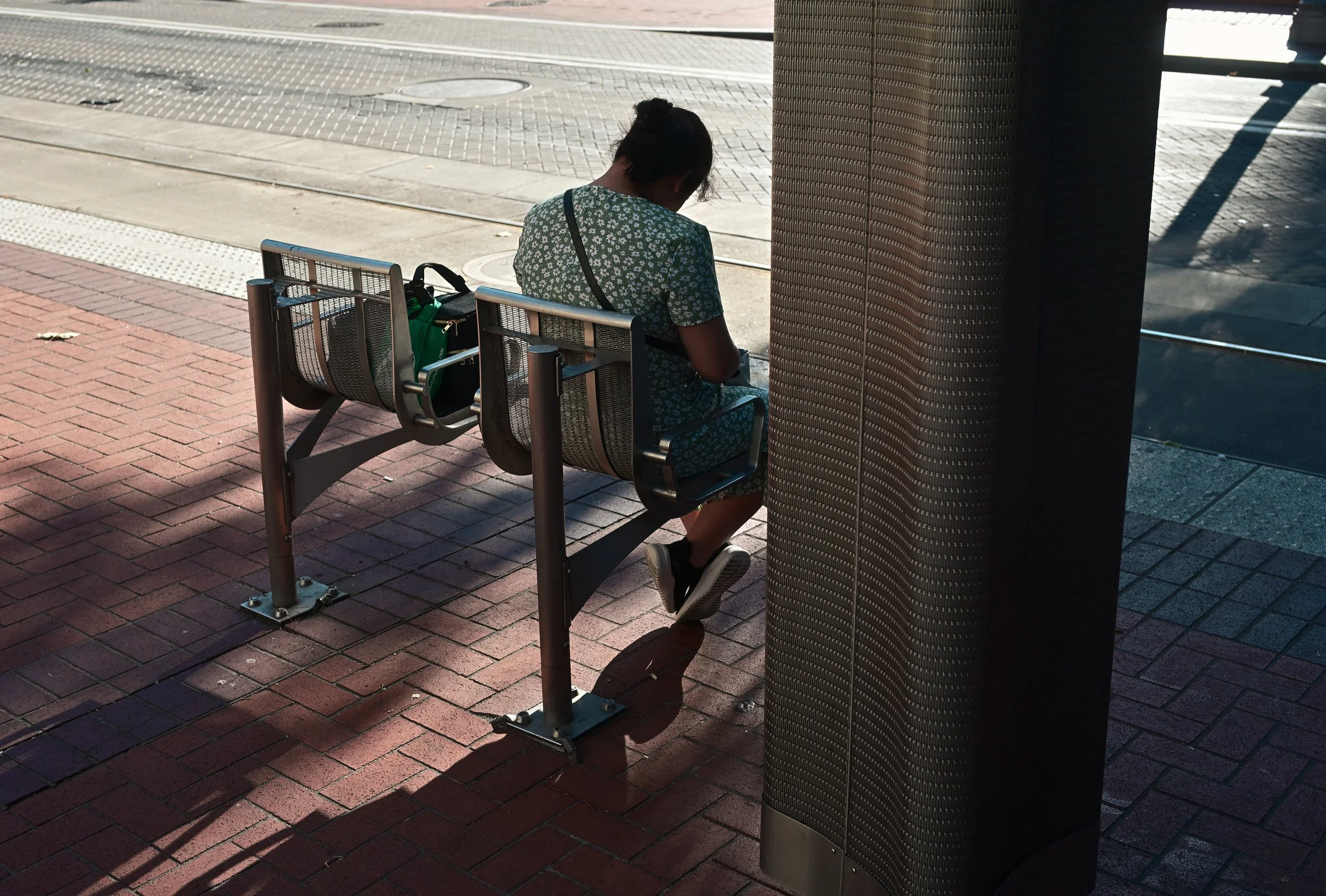 A woman sitting on a bus stop bench, looking at her phone, with a green bag placed beside her. She is wearing a green dress with white floral patterns. The background shows a sidewalk and street with tram tracks and shadows cast by nearby objects.