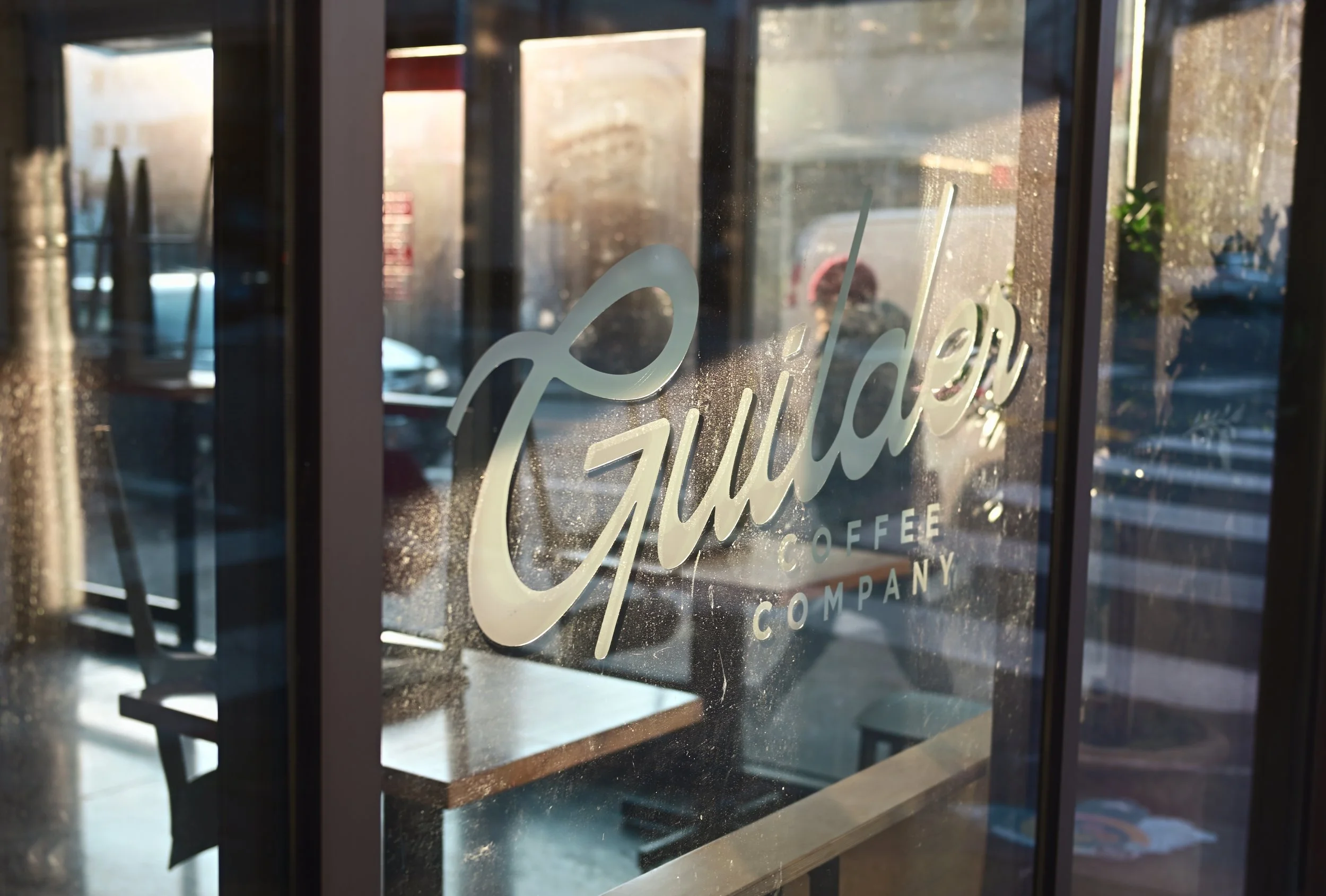 Glass window with the text 'Fulton' written on it, showing an interior space and some outdoor objects like a plant and parked cars, with sunlight casting reflections.