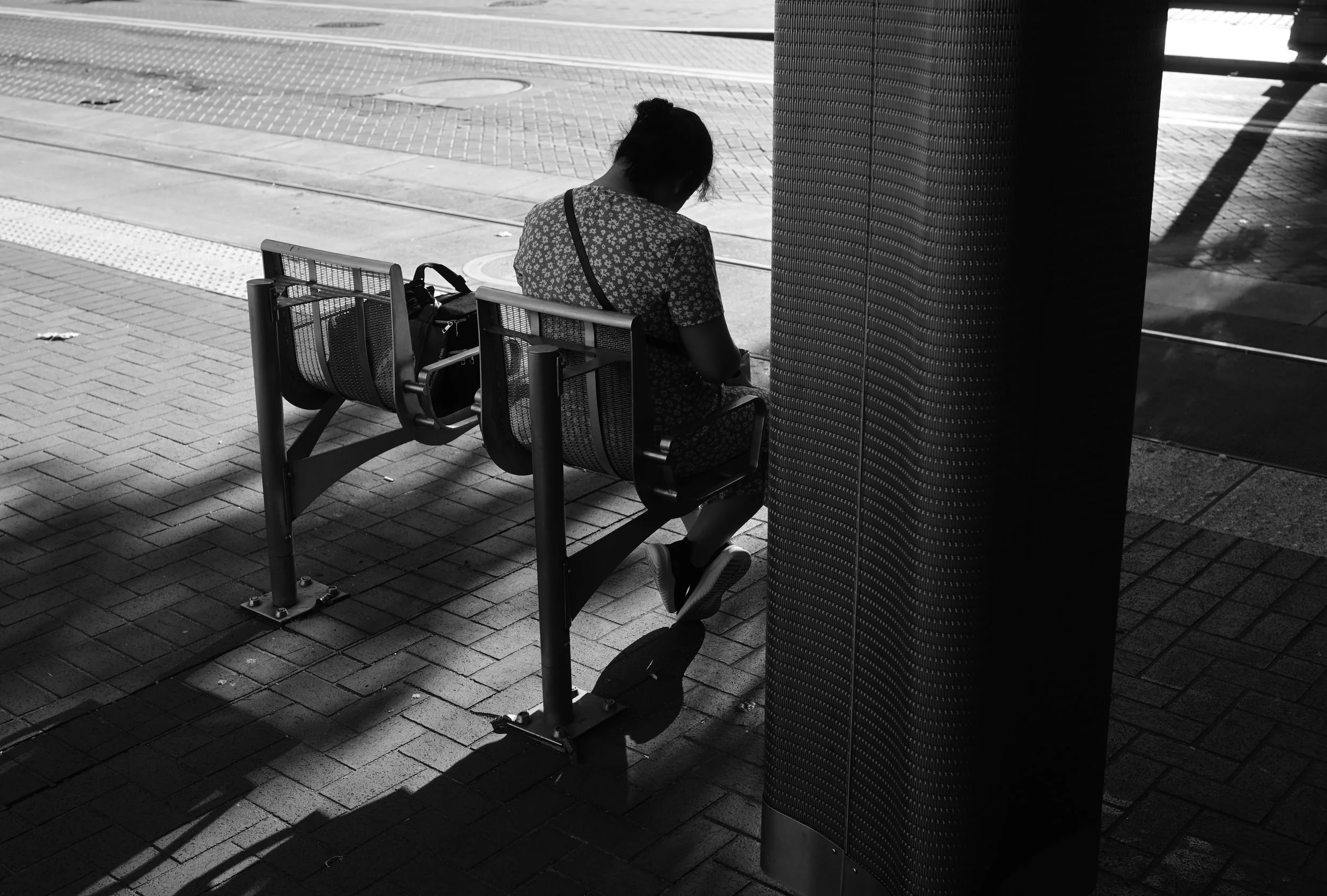 A person sitting alone on a bench at a bus stop, with their head bowed, in an urban setting, black and white photograph.