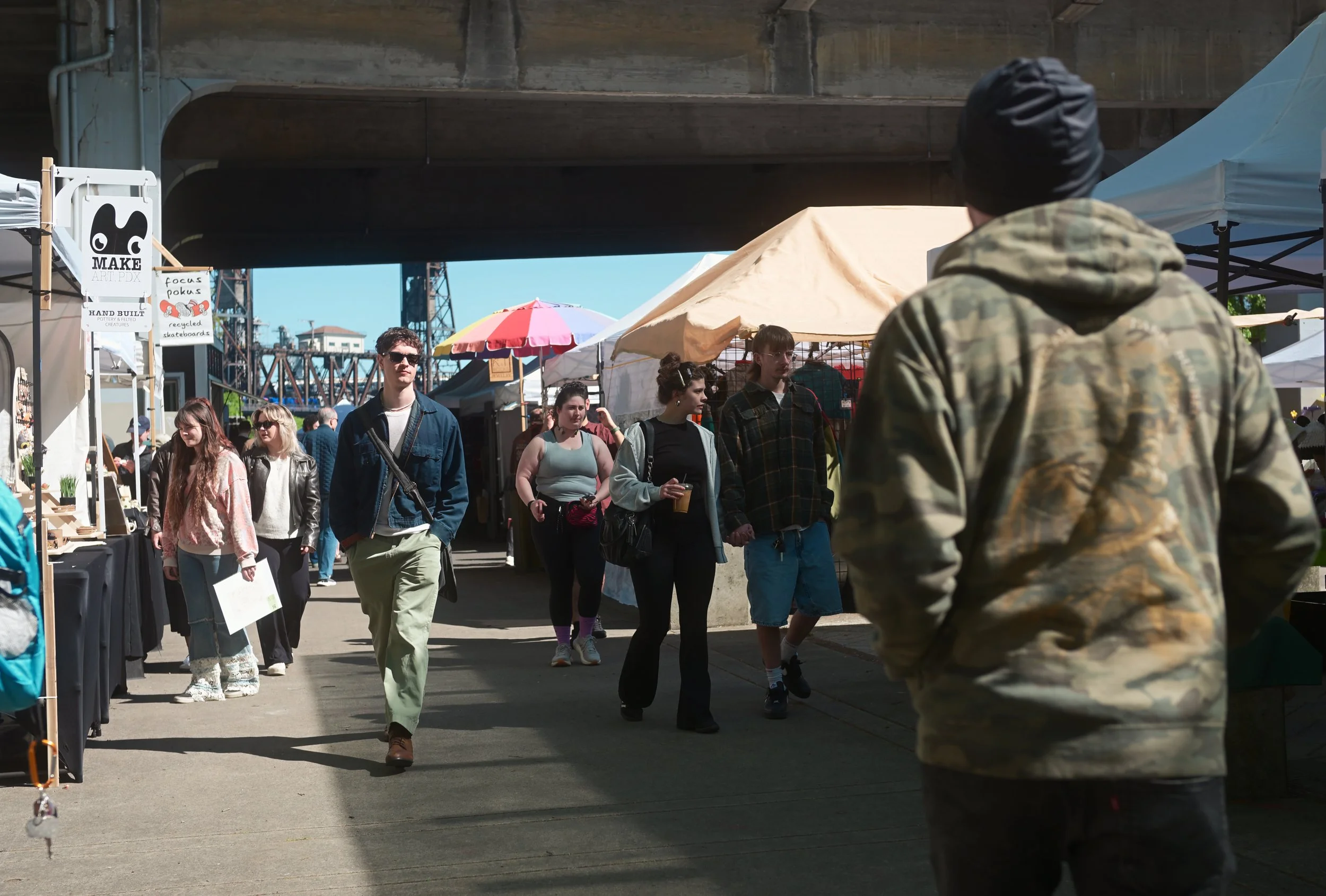 People walking through an outdoor market under a bridge. Stalls with tents and an umbrella are visible, and a man in camouflage clothing with his back to the camera is in the foreground.