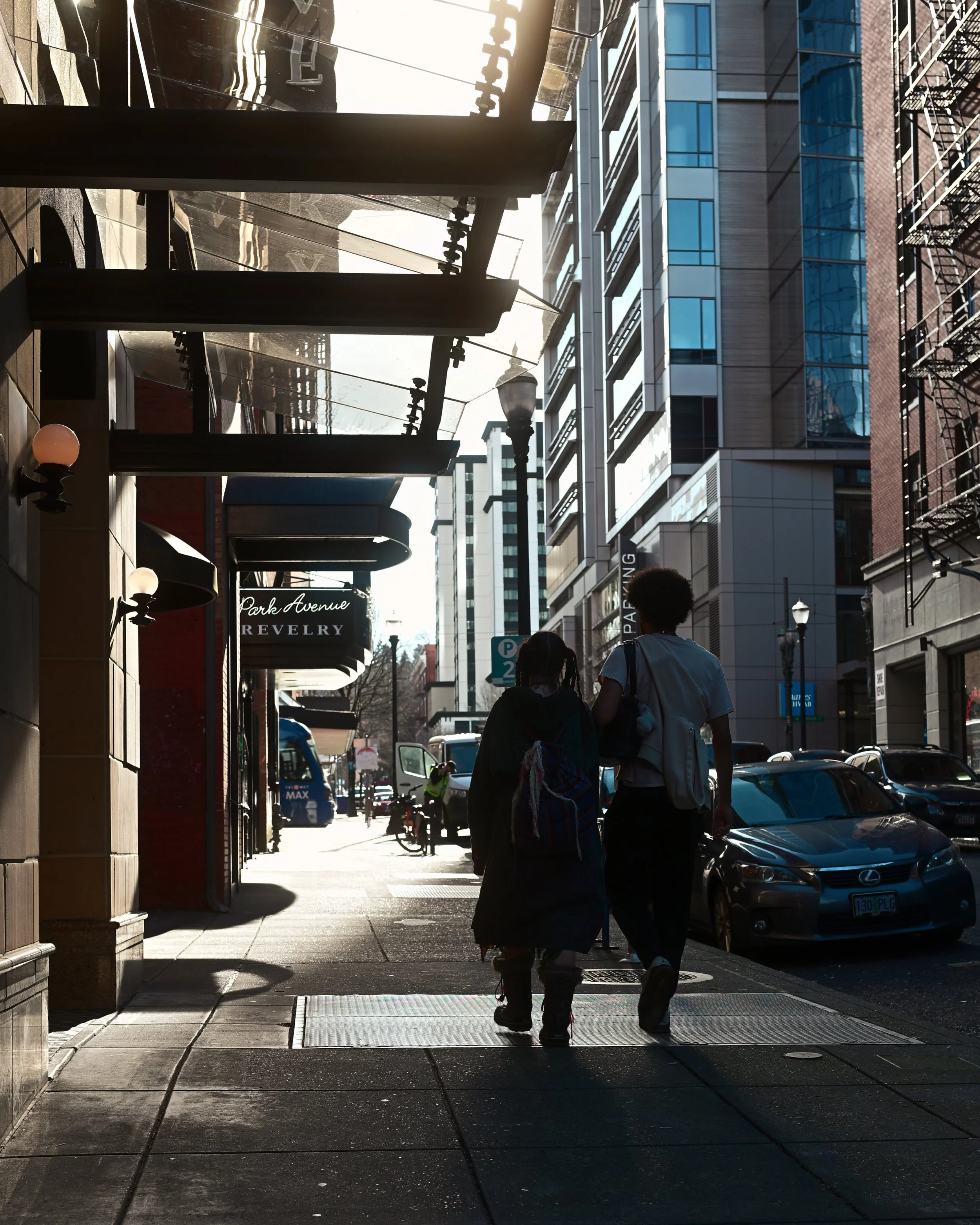 Two people walking on a city sidewalk with cars parked along the street and modern buildings in the background. The sun is shining brightly from behind the buildings, creating shadows and reflections.
