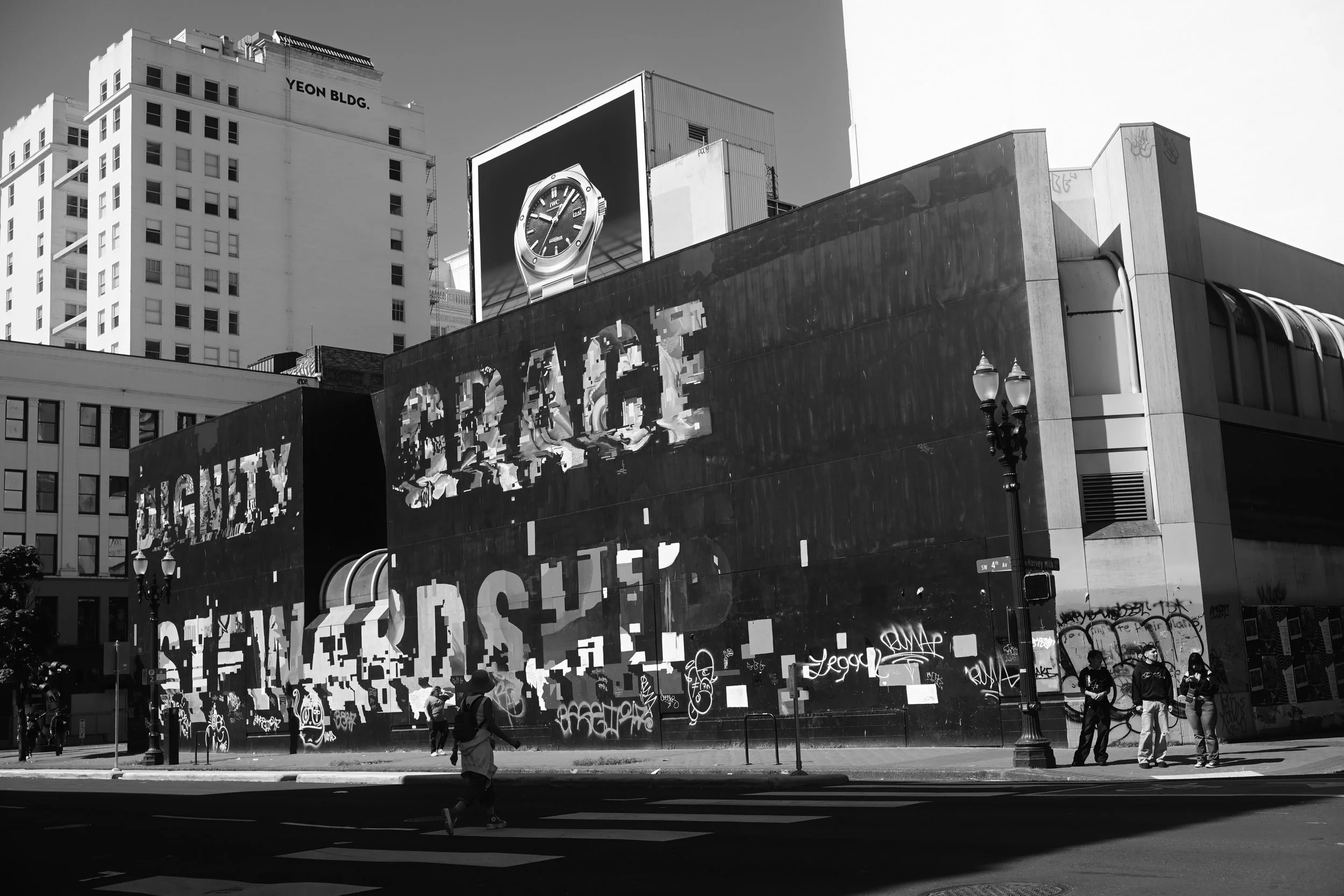 Black and white photo of an urban street scene featuring a large building with multiple stories and windows, with a billboard advertising a watch. There are several people on the sidewalk, some wearing masks, and graffiti on the lower part of a wall. A vintage-style streetlamp is also visible.