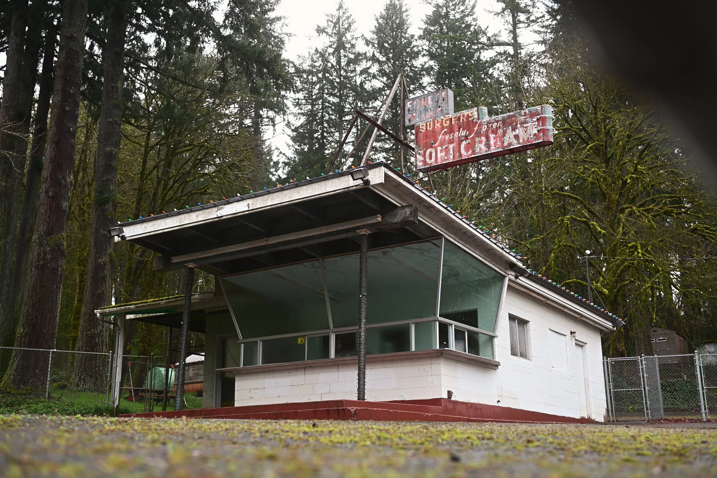 A vintage roadside burger stand with a faded red sign reading "Burgers, Soft Serve" in a wooded area, surrounded by tall trees and moss.