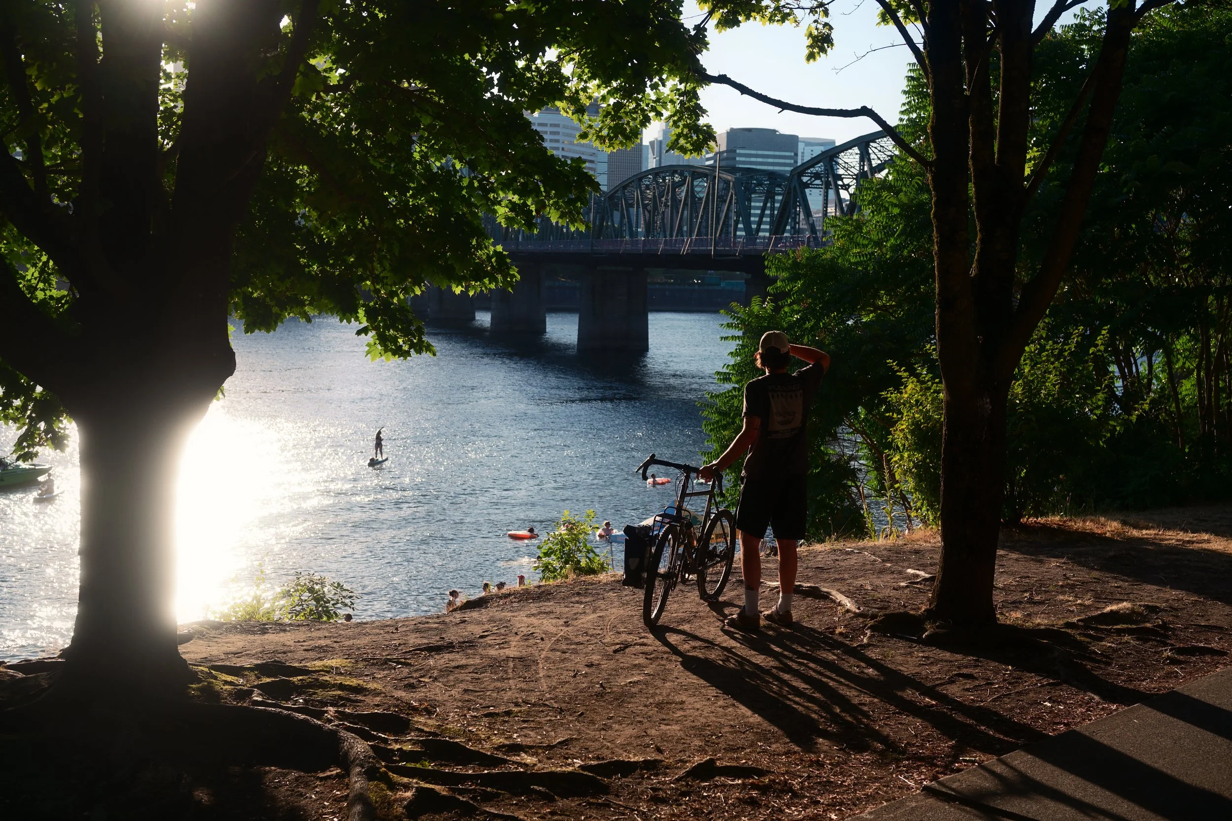 A man with a bicycle on a riverside, shaded by trees, with a bridge and cityscape in the background, and people swimming and paddleboarding on the water.