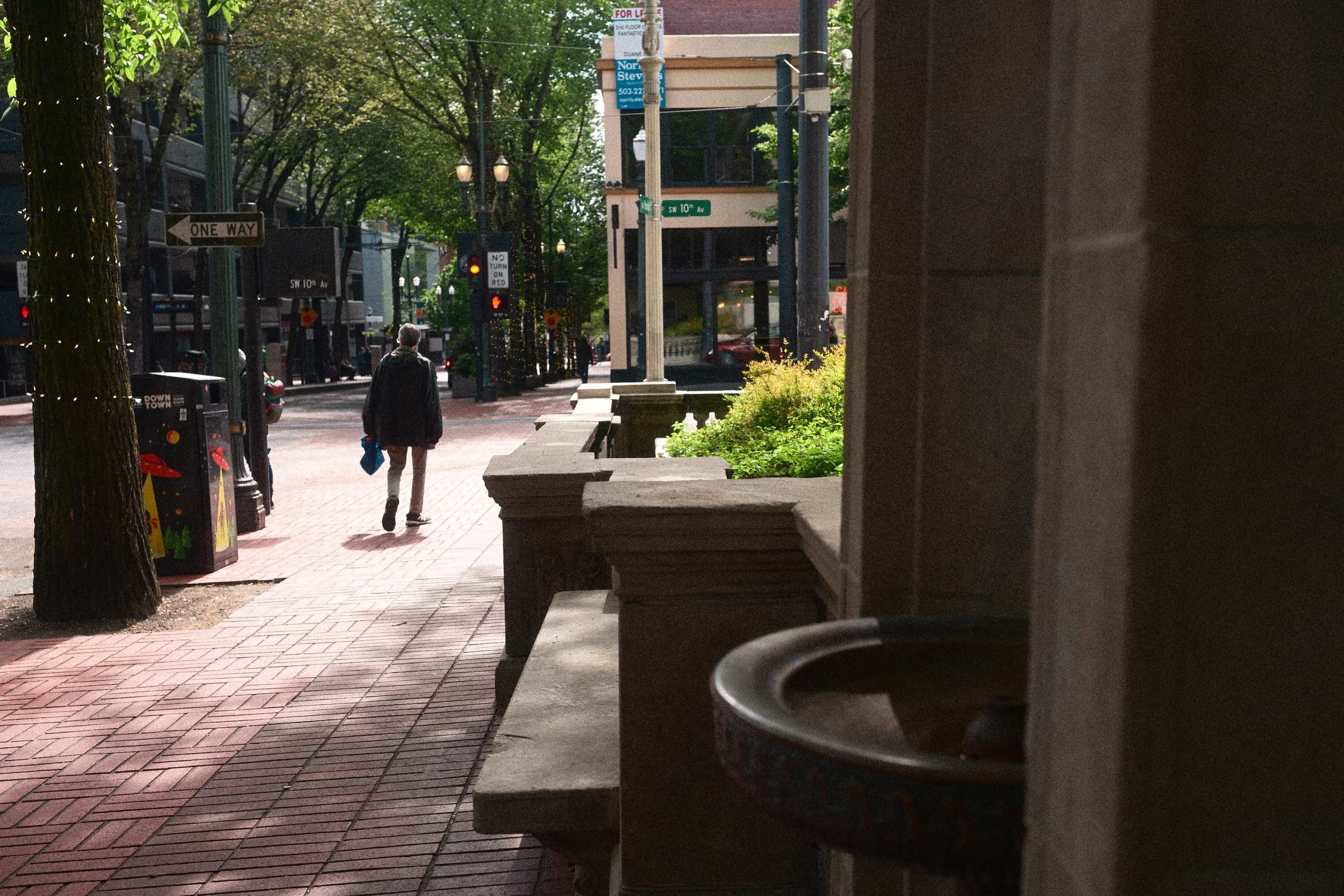 Pedestrian walking along a brick-paved downtown Portland sidewalk at SW 10th Avenue, beneath tree-lined streets and street signs.