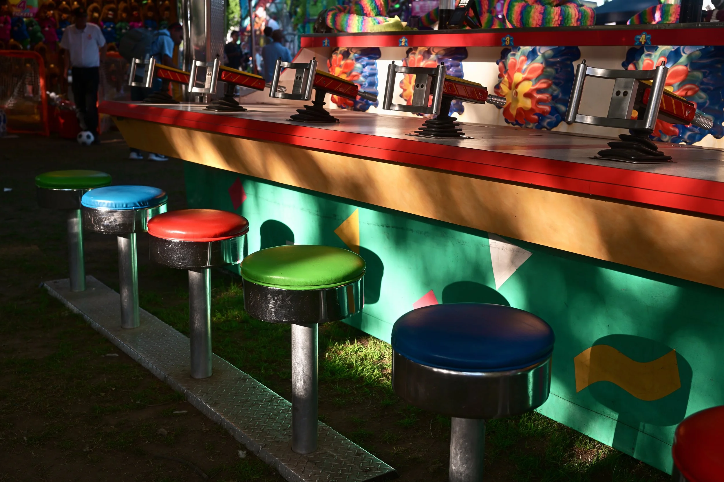 Colorful carnival game booth with round stools in red, green, blue, and black in front, featuring a wall of stuffed animals and prizes behind.