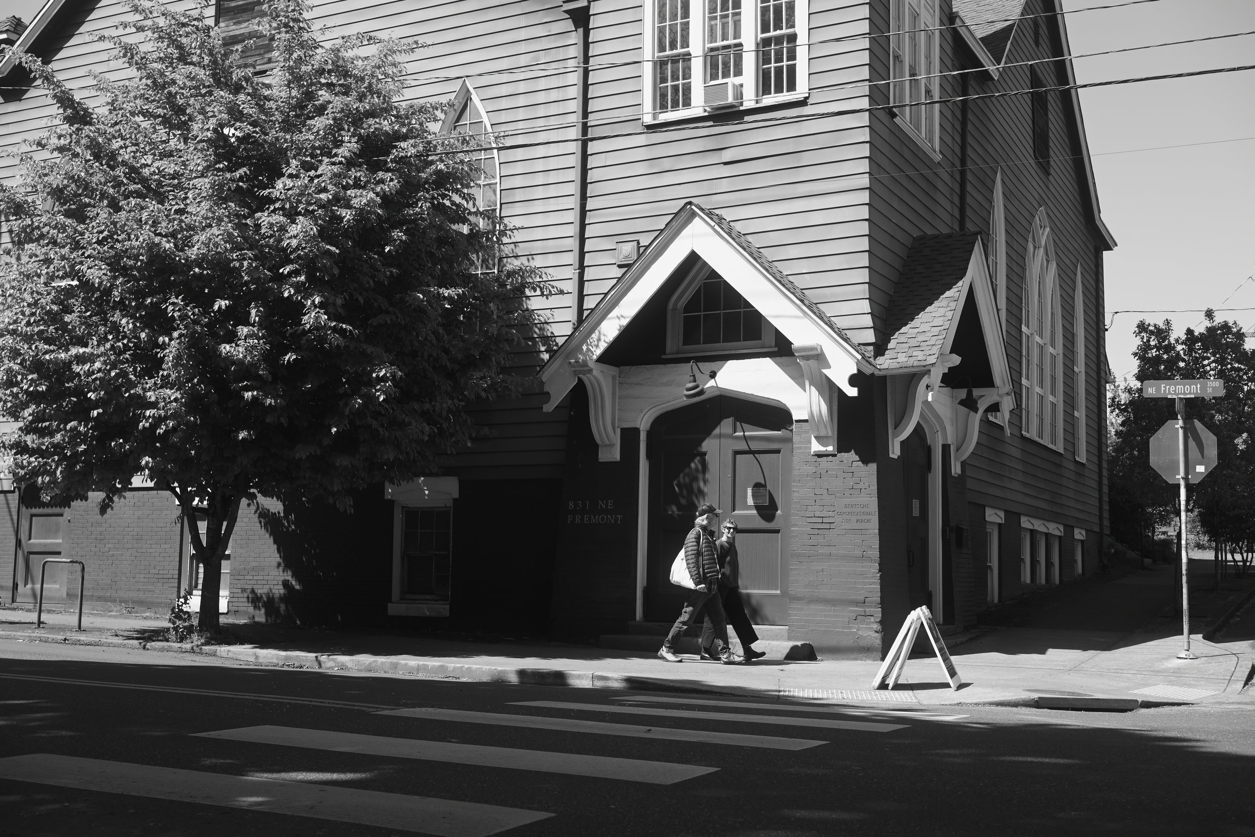 Black and white photo of a street corner with a large wooden house, two people walking on the sidewalk, and street signs that read 'NE Fremont' and '3500'.