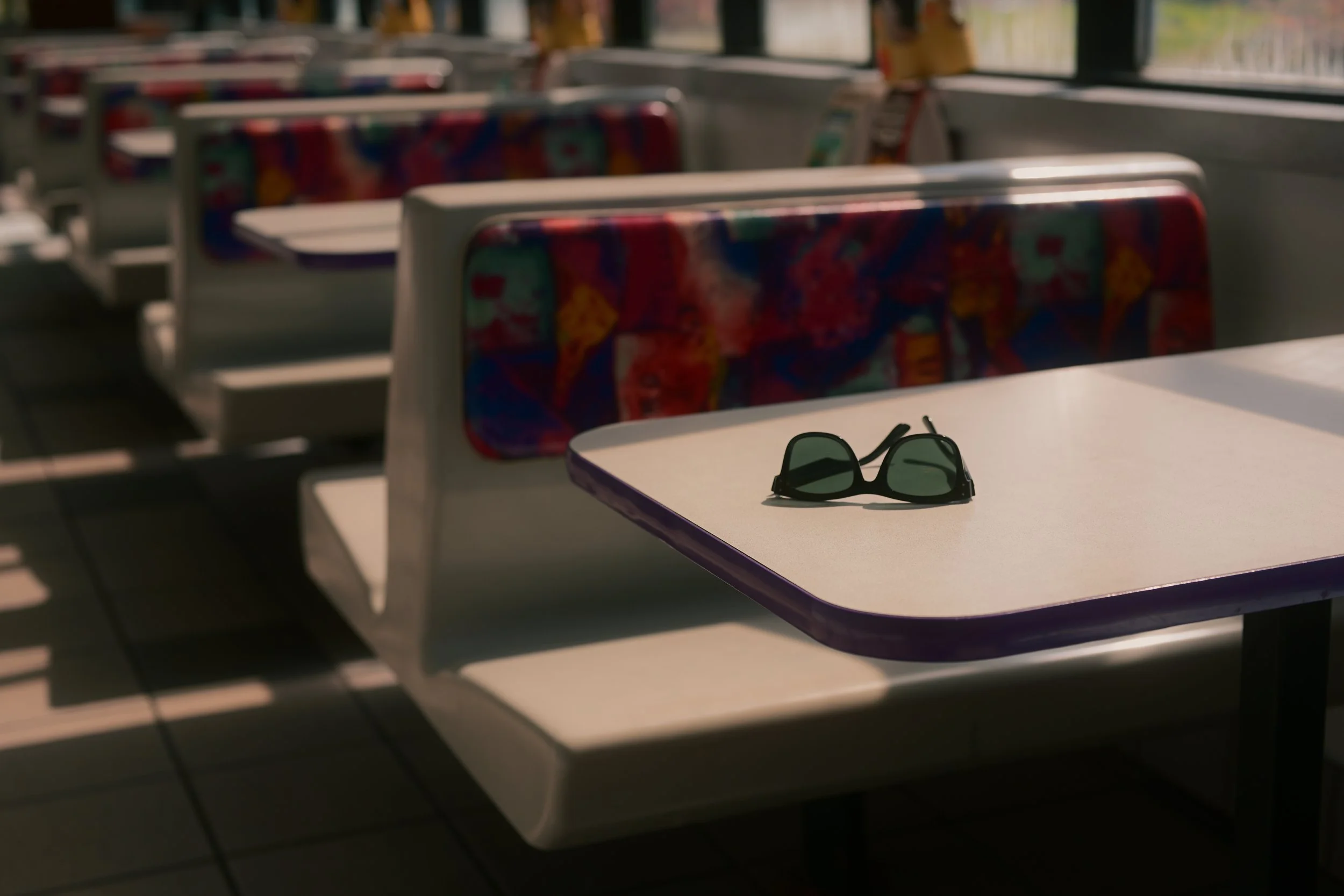 A pair of black sunglasses on a white table near a window in a train or bus, with rows of colorful patterned seats in the background.