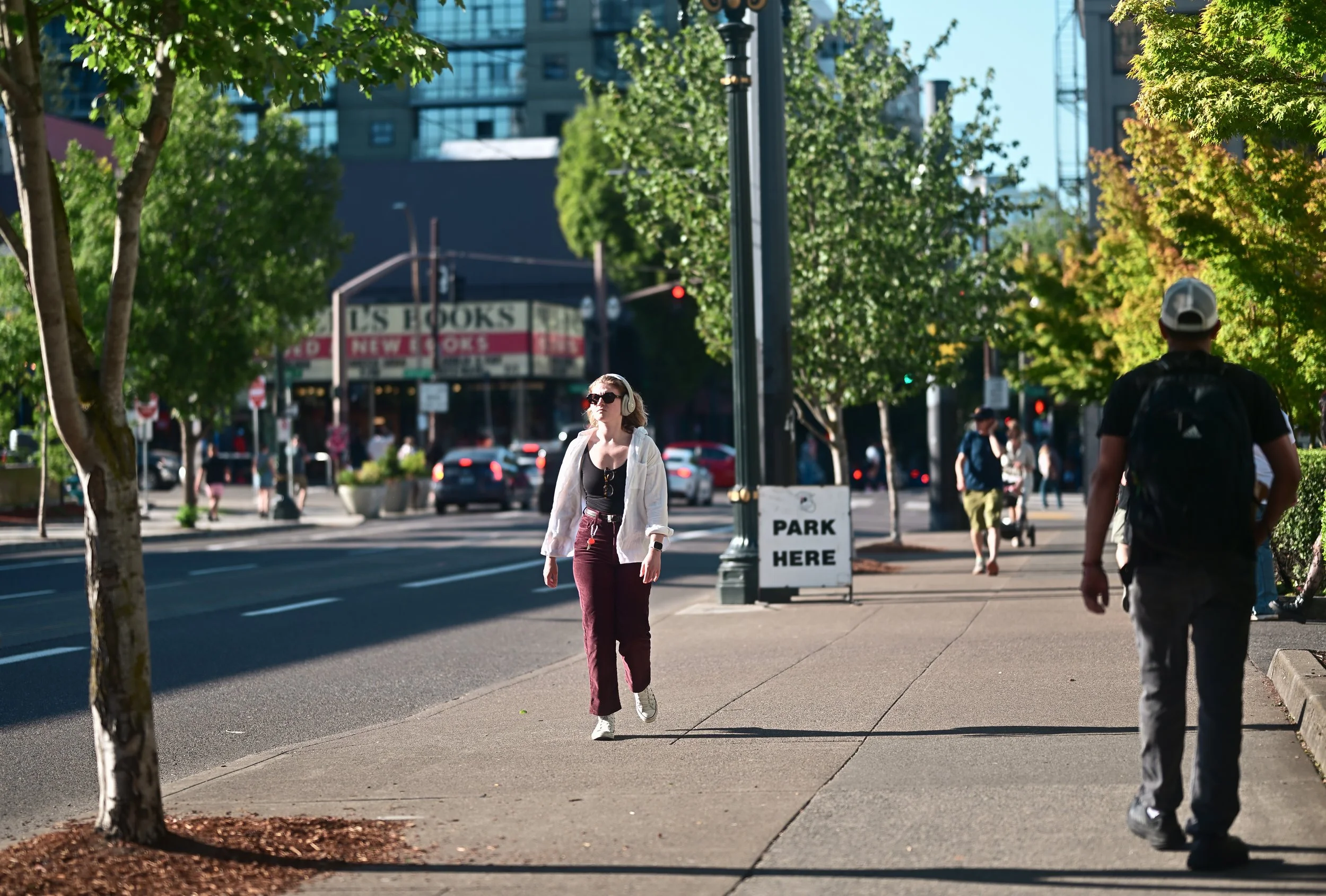 A woman with headphones walking on a city sidewalk lined with trees, other pedestrians, and parked cars, with tall buildings and a store sign in the background.