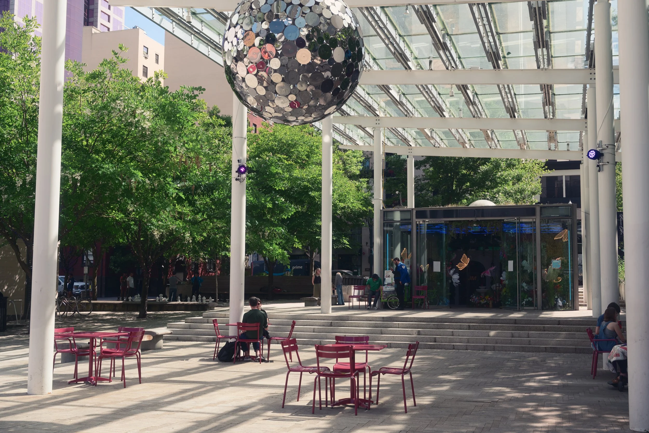 An outdoor plaza with pink chairs and tables, a large disco ball hanging from a glass and metal roof structure, trees providing shade, and a glass display case with butterfly decorations. People are sitting, walking, and talking in the area.