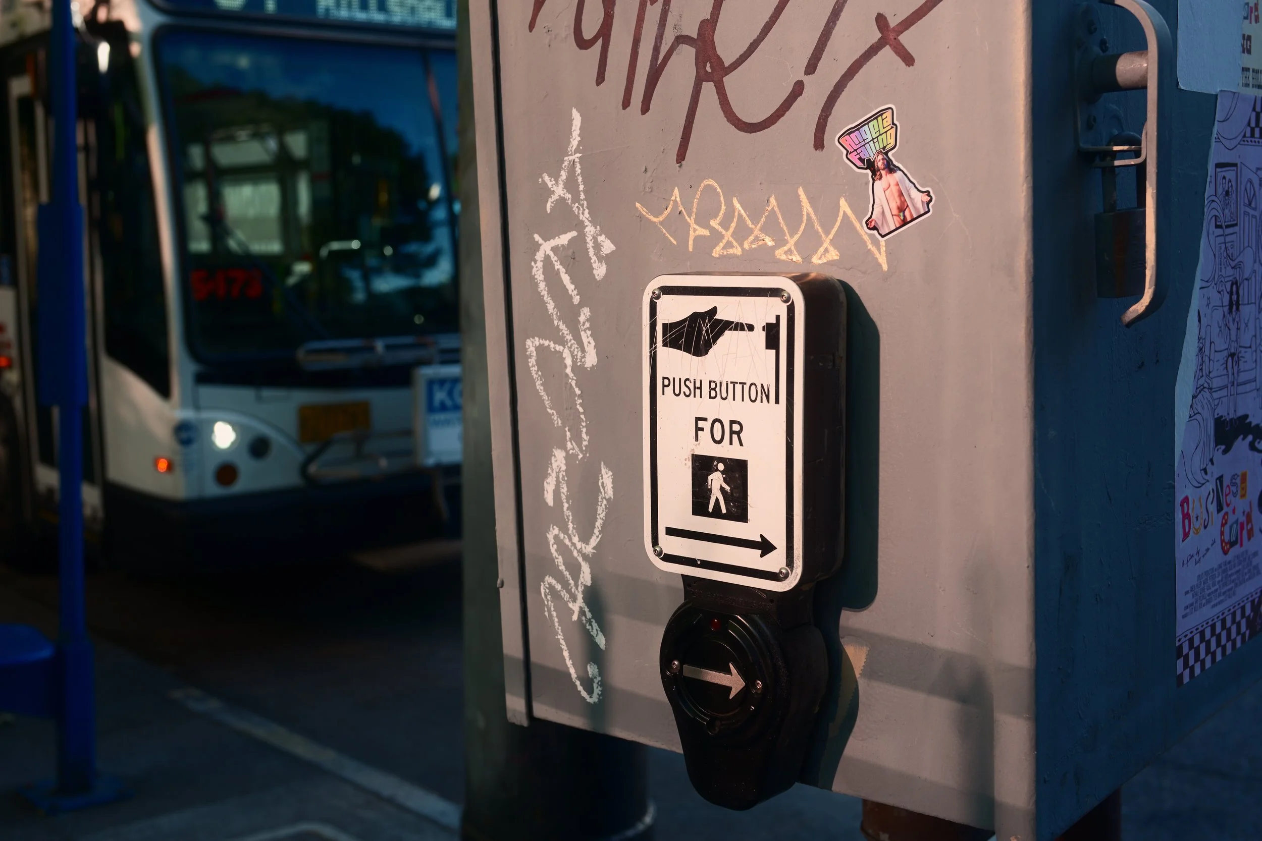 Pedestrian crossing button with an arrow pointing right, mounted on a pole near a bus at a bus stop. The background includes a bus and some street signs, with graffiti and stickers on the pole.